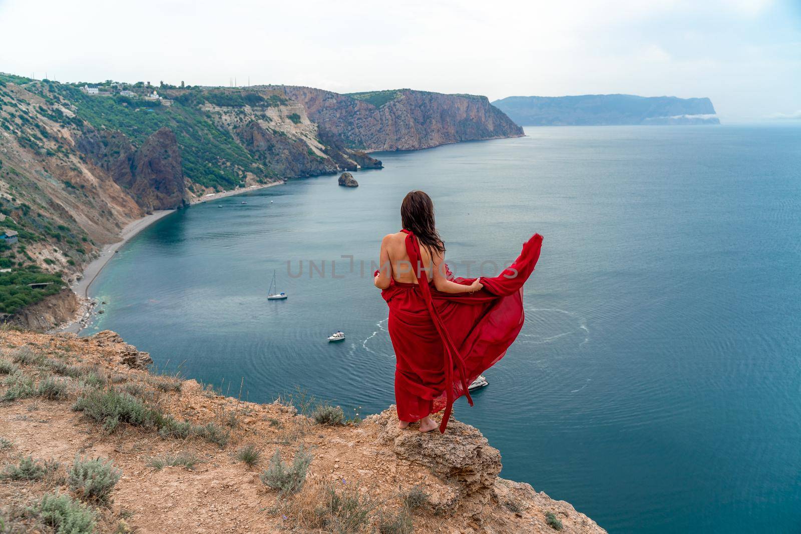 A girl with loose hair in a long red dress descends the stairs between the yellow rocks overlooking the sea. A rock can be seen in the sea. Sunny path on the sea from the rising sun by Matiunina