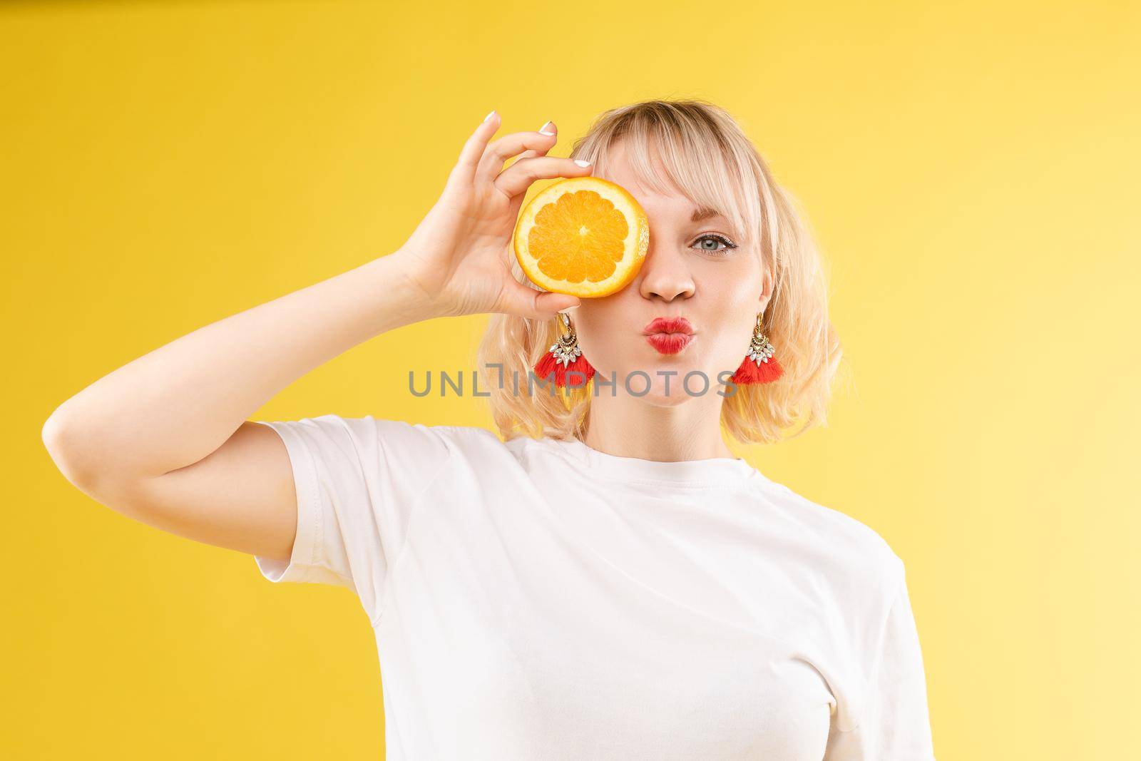 Stock photo of positive blonde young woman in white t-shirt with halved orange holding it in front of her eye and pouting lips at camera. Isolate on yellow background. Summer concept.