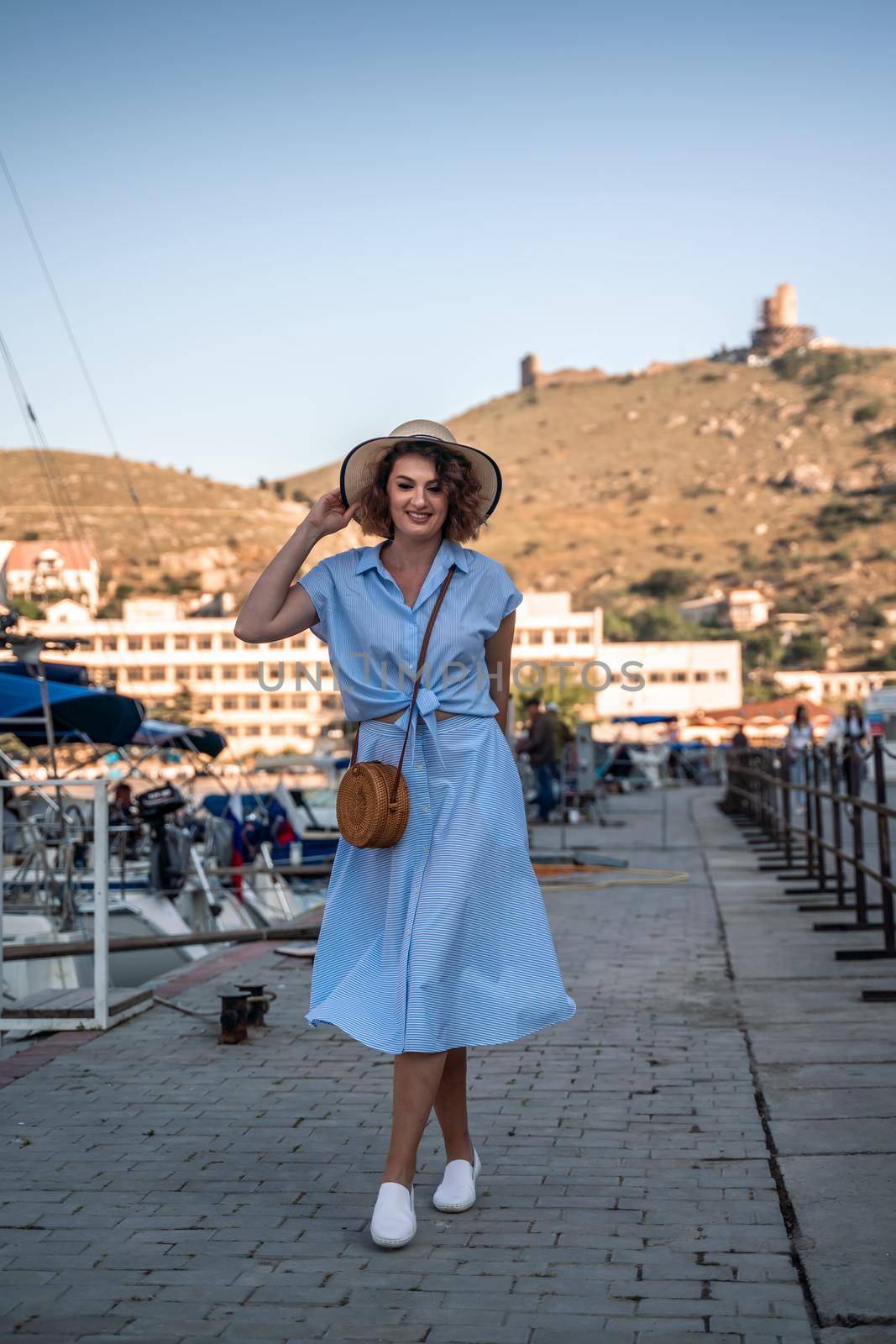 A young happy woman in a blue dress and hat stands near the seaport with luxury yachts. Travel and vacation concept by Matiunina