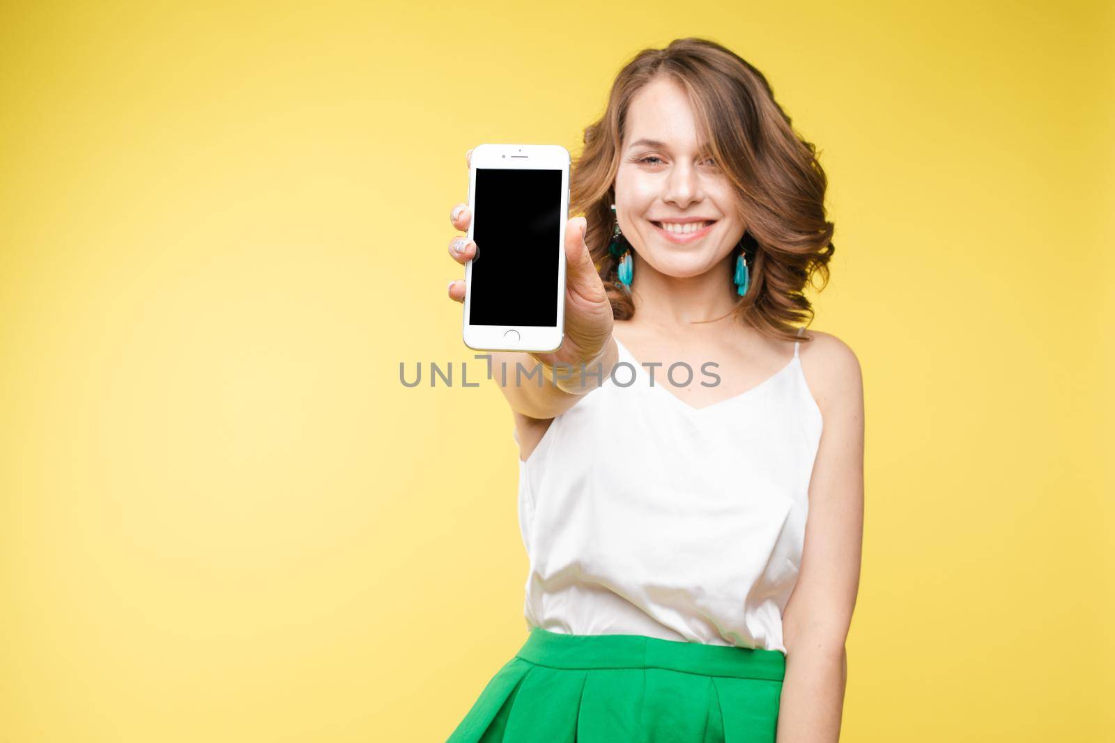 Studio portrait of amazed young caucasian woman with wavy hair wearing white top and green bottom holding mobile phone screen to camera. She is shocked or surprised with the news or info on the screen. Copyspace. Isolate on yellow.