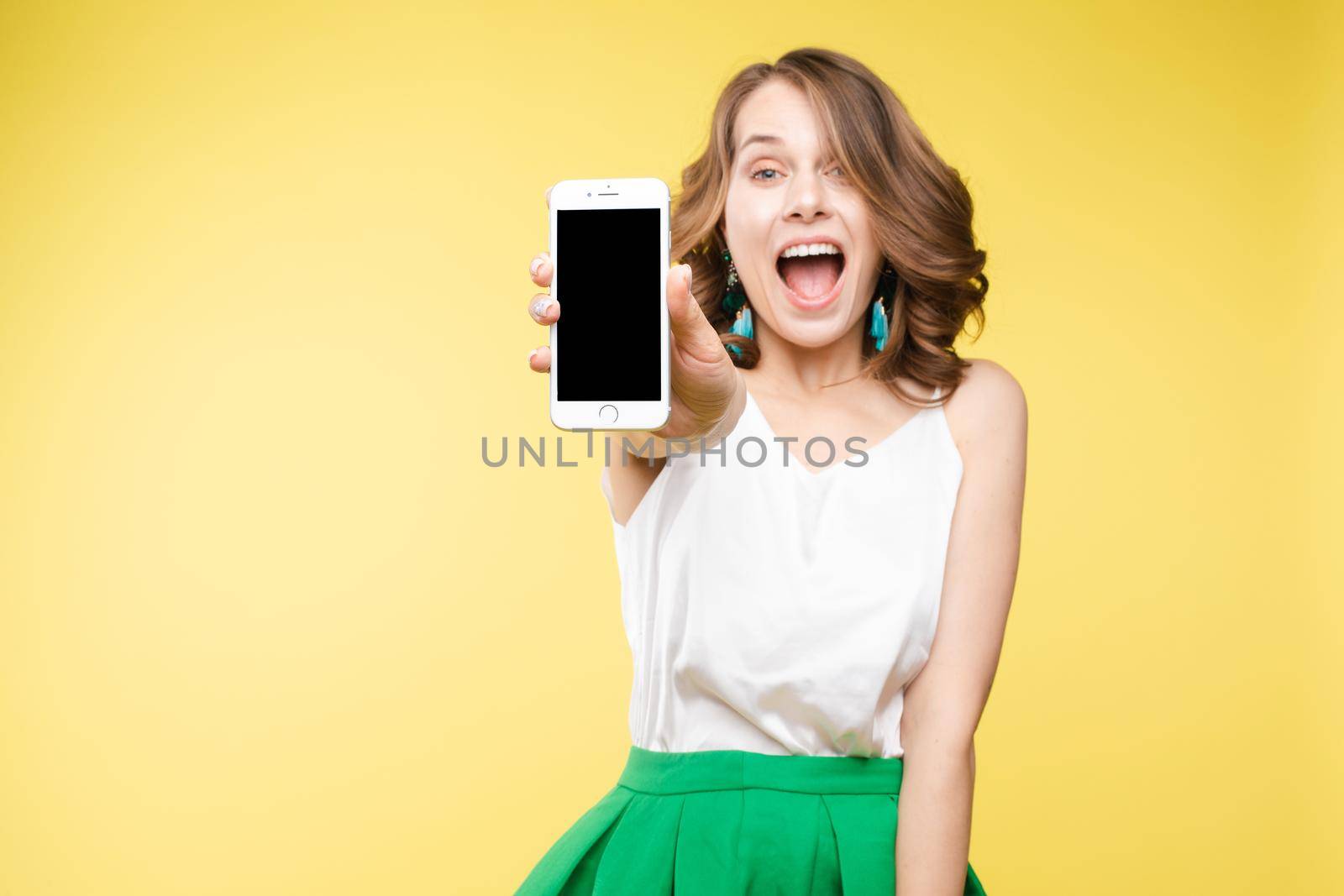 Studio portrait of amazed young caucasian woman with wavy hair wearing white top and green bottom holding mobile phone screen to camera. She is shocked or surprised with the news or info on the screen. Copyspace. Isolate on yellow.