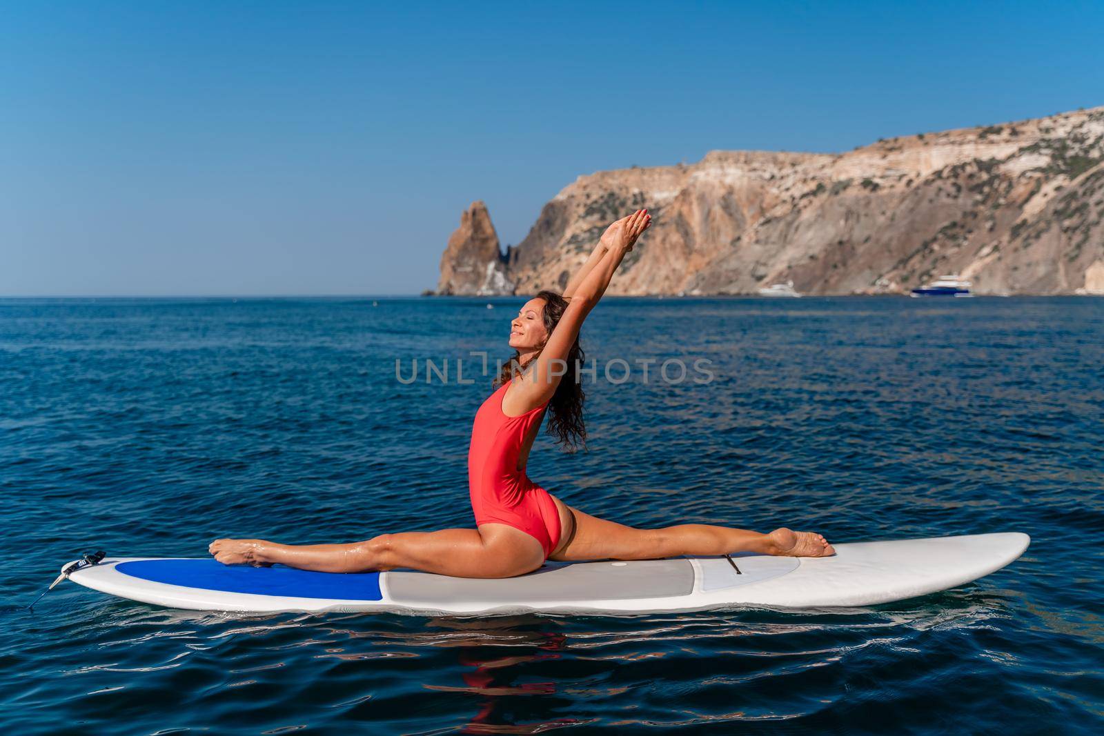 Sporty girl on a surfboard in the sea on a sunny summer day. In a red swimsuit, she sits in the splits on the sap. Summer entertainment on Stortom by the sea.