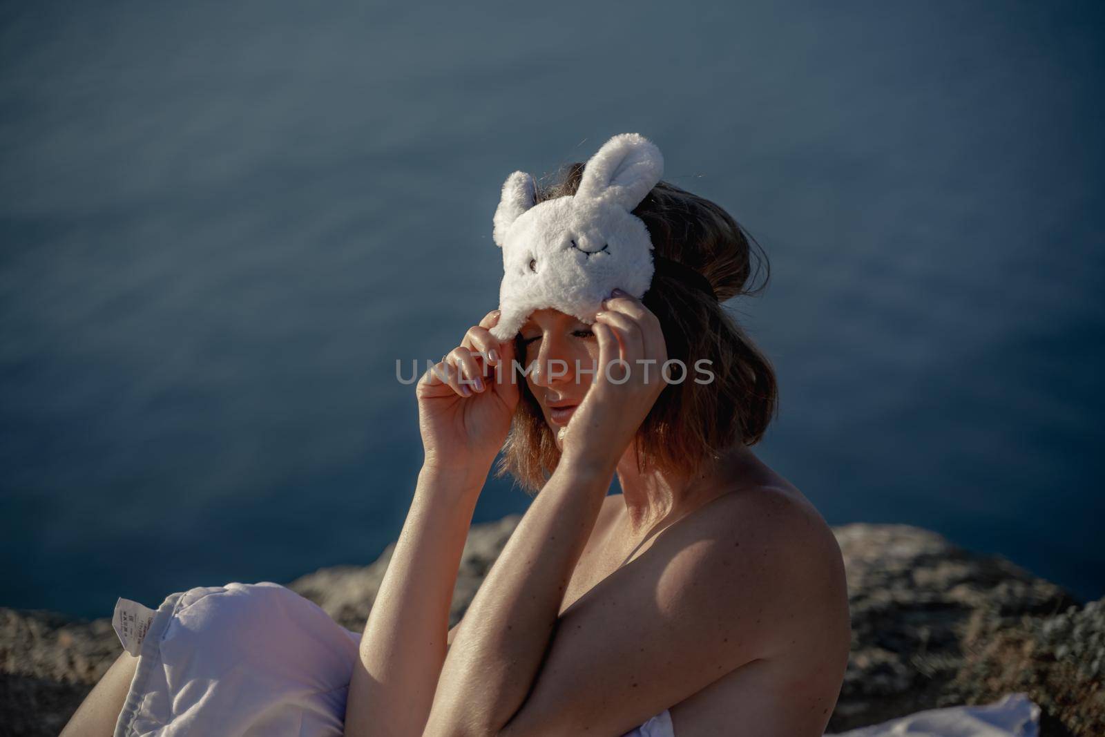 Young woman in sleep mask posing on the seashore. She is wrapped in a white blanket. The coast of the sea at a height, around the sea and rocks, dawn. by Matiunina