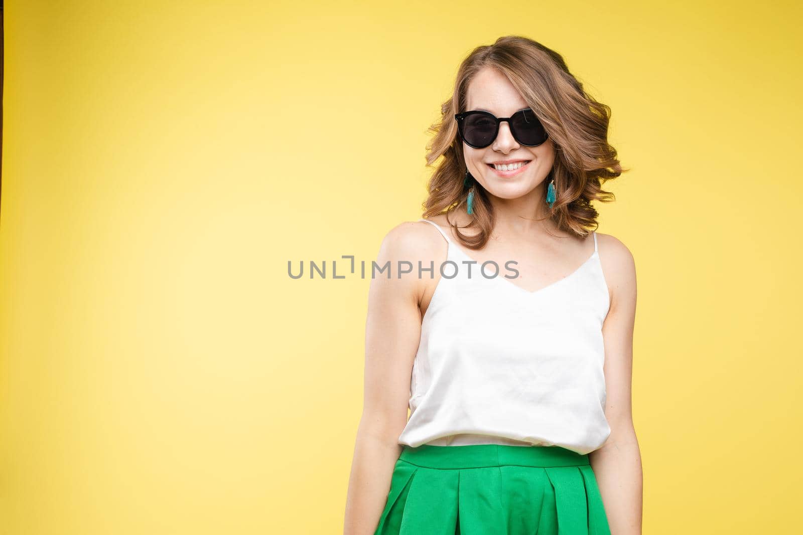 Summer outfit on orange background. Stylish and trendy beautiful young girl with wavy middle-length hair in white top and green bottom wearing trendy earrings and black sunglasses. She is smiling over orange background. Copyspace. Isolate.