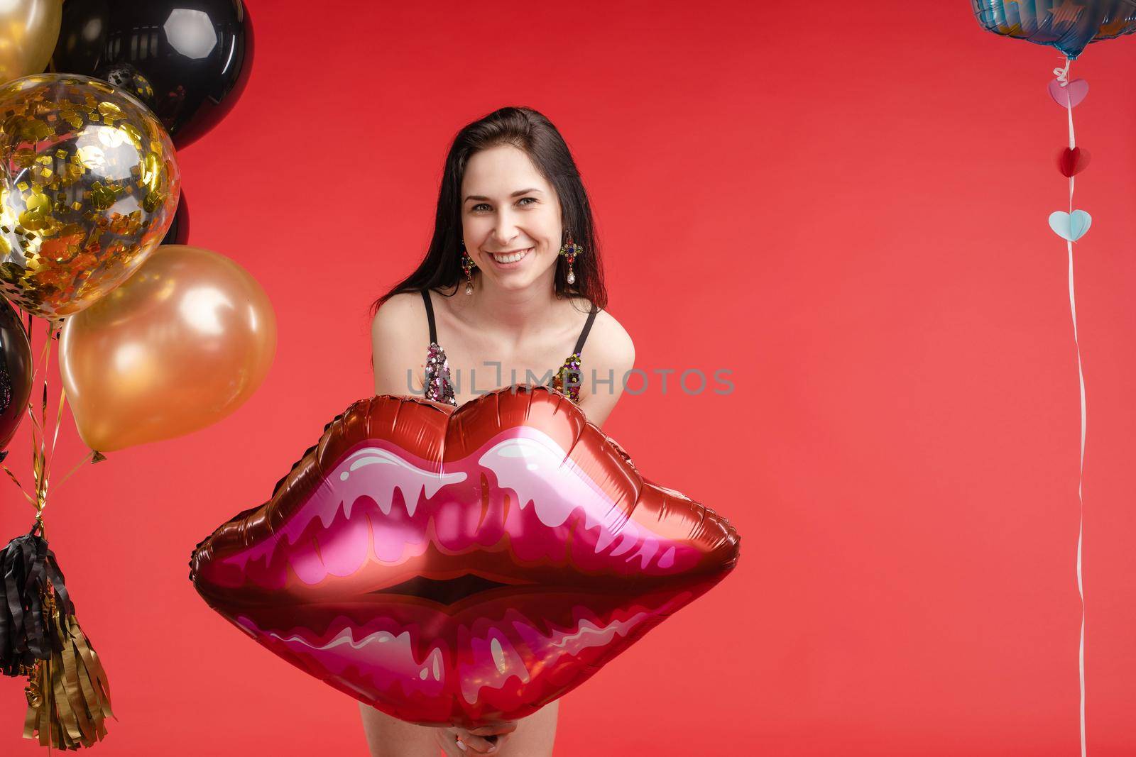 Front view of cheerful attractive girl in bright short dress dancing and laughing on red isolated background. Happy brunette posing near balloons in studio. Smiling woman celebrating new year party.