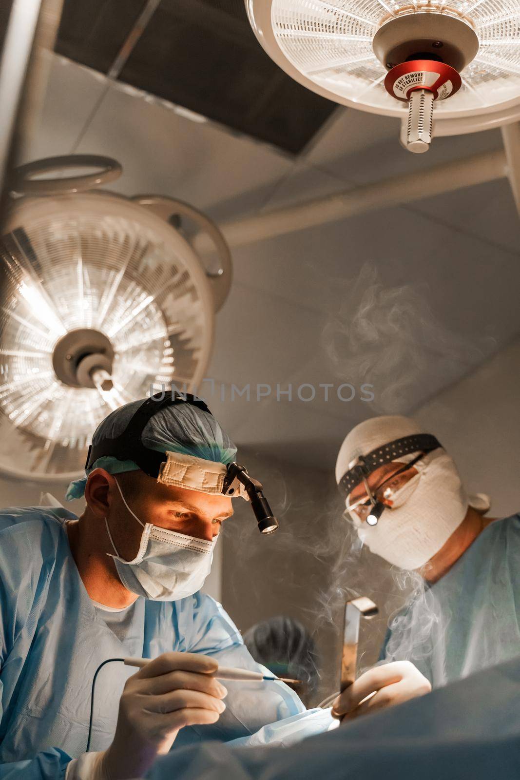 2 surgeons with headlamp do plastic operation in medical clinic. Chest augmentation plastic operation and correction in medical clinic. by Rabizo