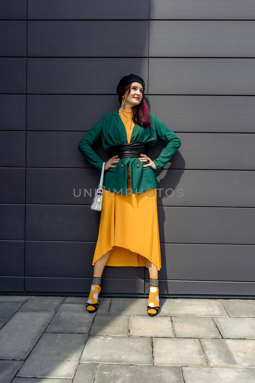 Young woman in bright clothes, yellow skirt and green jacket. Yellow socks in sandals, beret on the head, hair with the color of magenta. Caucasian female fashion model standing against gray wall background, open with copy space. by Matiunina