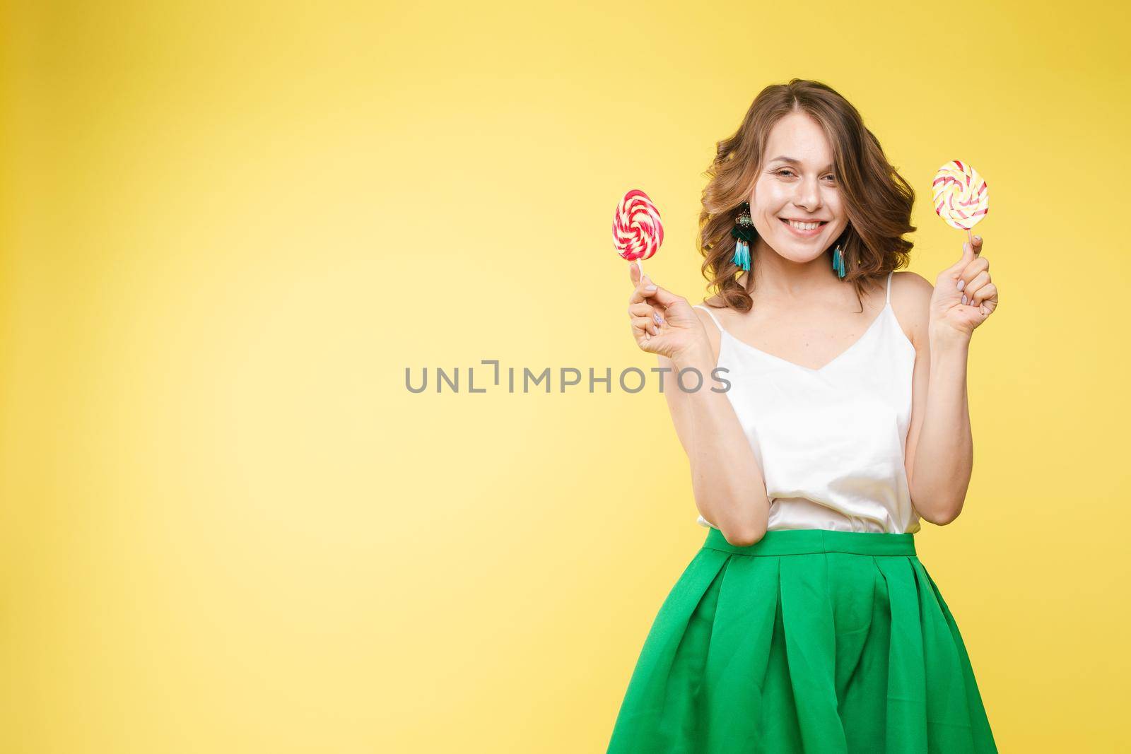 Full length studio portrait of laughing model in white top and green skirt and heels holding two sweet candies on her eyes like sunglasses with her mouth open. Isolate on yellow.