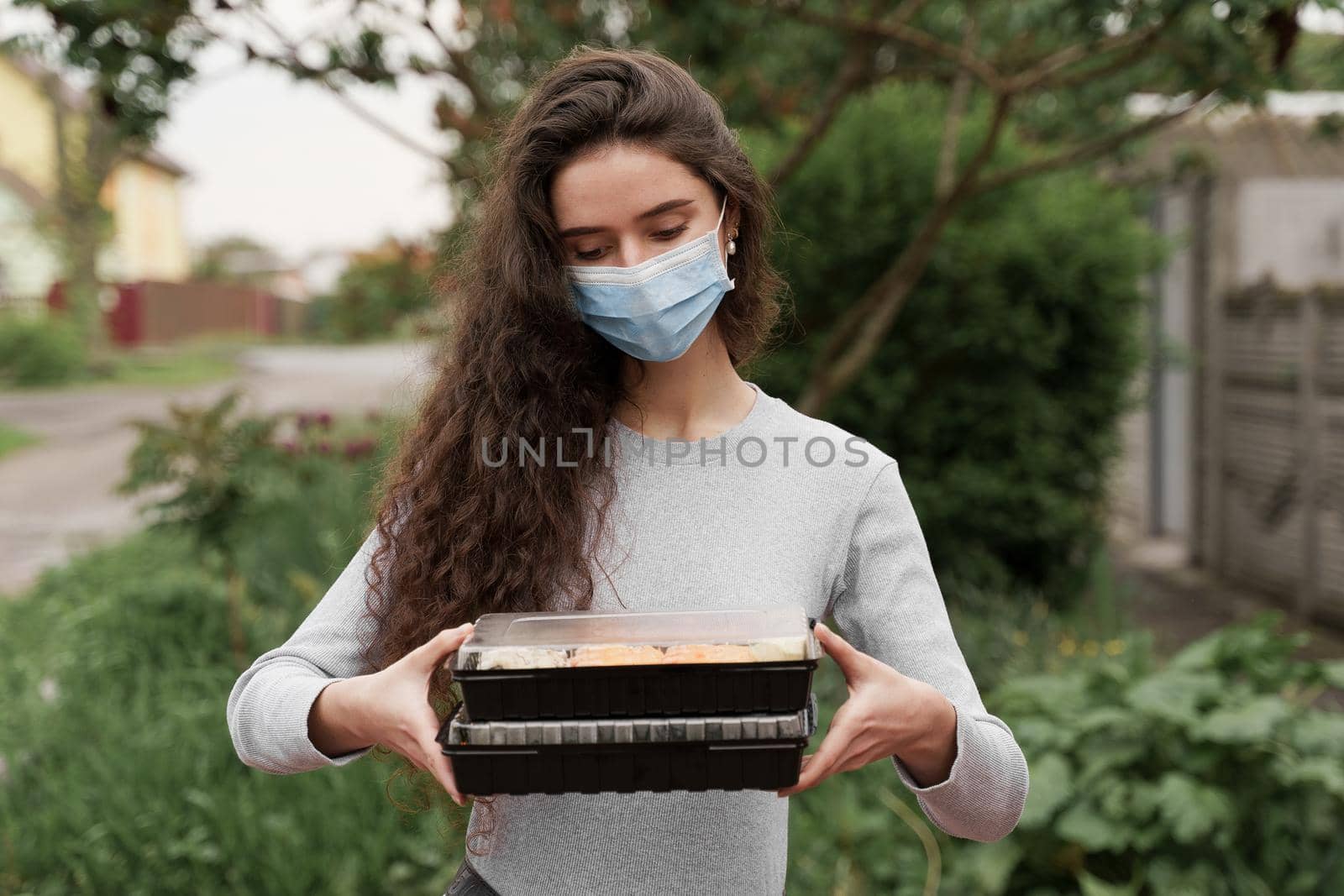 Girl courier in medical mask with 2 sushi boxes stands in front of car. Sushi set in box healthy food delivery service by car