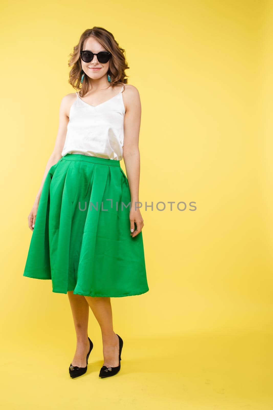 Summer outfit on orange background. Stylish and trendy beautiful young girl with wavy middle-length hair in white top and green bottom wearing trendy earrings and black sunglasses. She is smiling over orange background. Copyspace. Isolate.