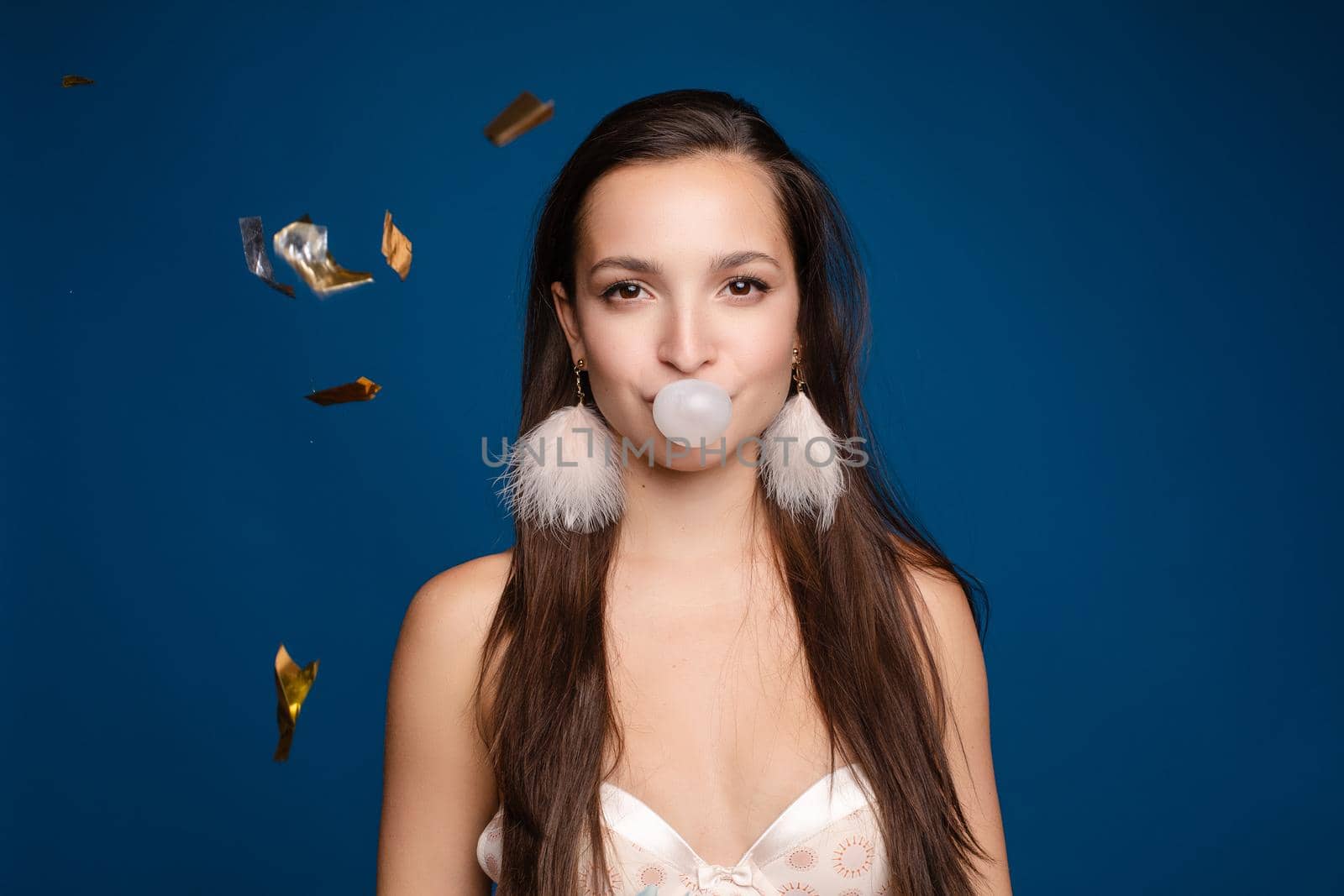 Front view of stylish female teenager wearing tracksuit and hat chewing gum and keeping big blue balloon in studio. Young girl looking at camera and posing on grey isolated background.