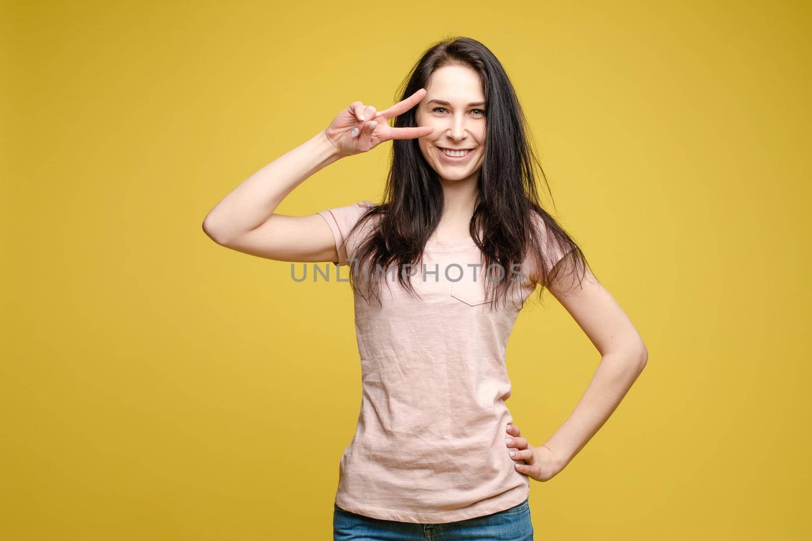 Studio portrait of charming brunette young woman in white top and green skirt holding peace sign near her eye. She is smiling at camera. Isolate on orange.
