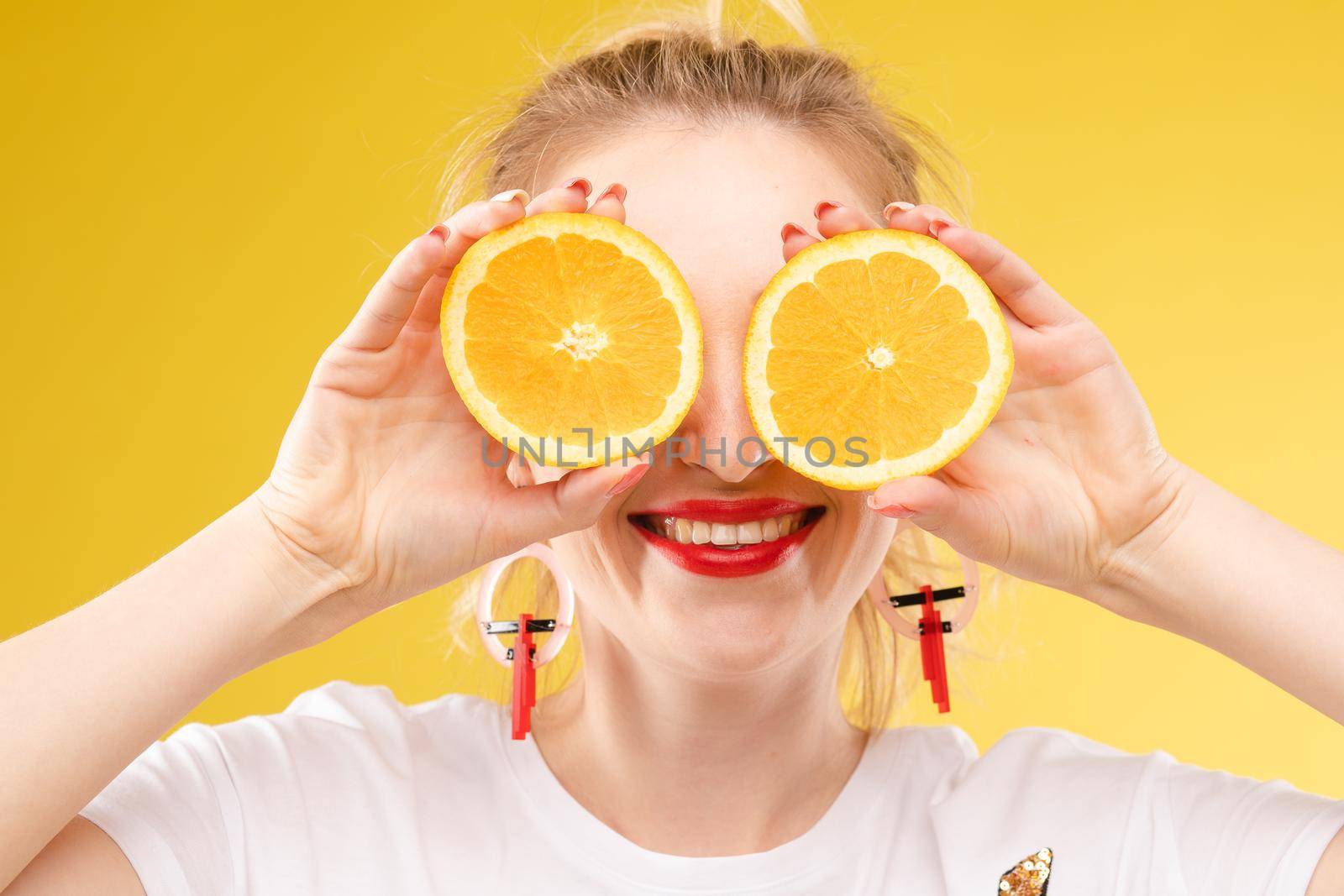 Front view of cheerful young blonde posing with fresh oranges on isolated background. Funny girl keeping fruit and closing eyes in studio. Concept of happiness and health.