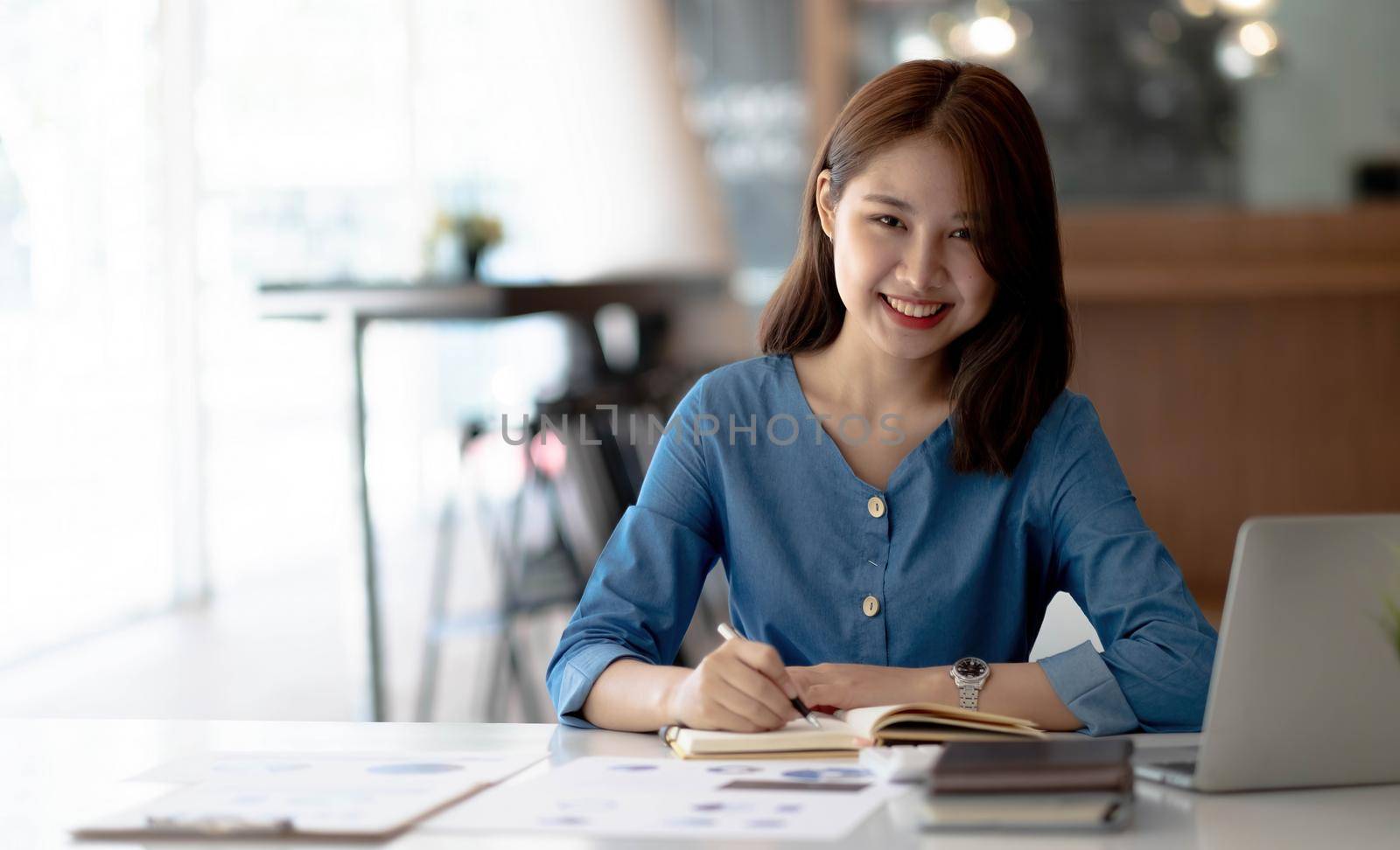 Cheerful young woman with laptop and jar and writing in notebook while sitting at table and working on freelance project at home by wichayada