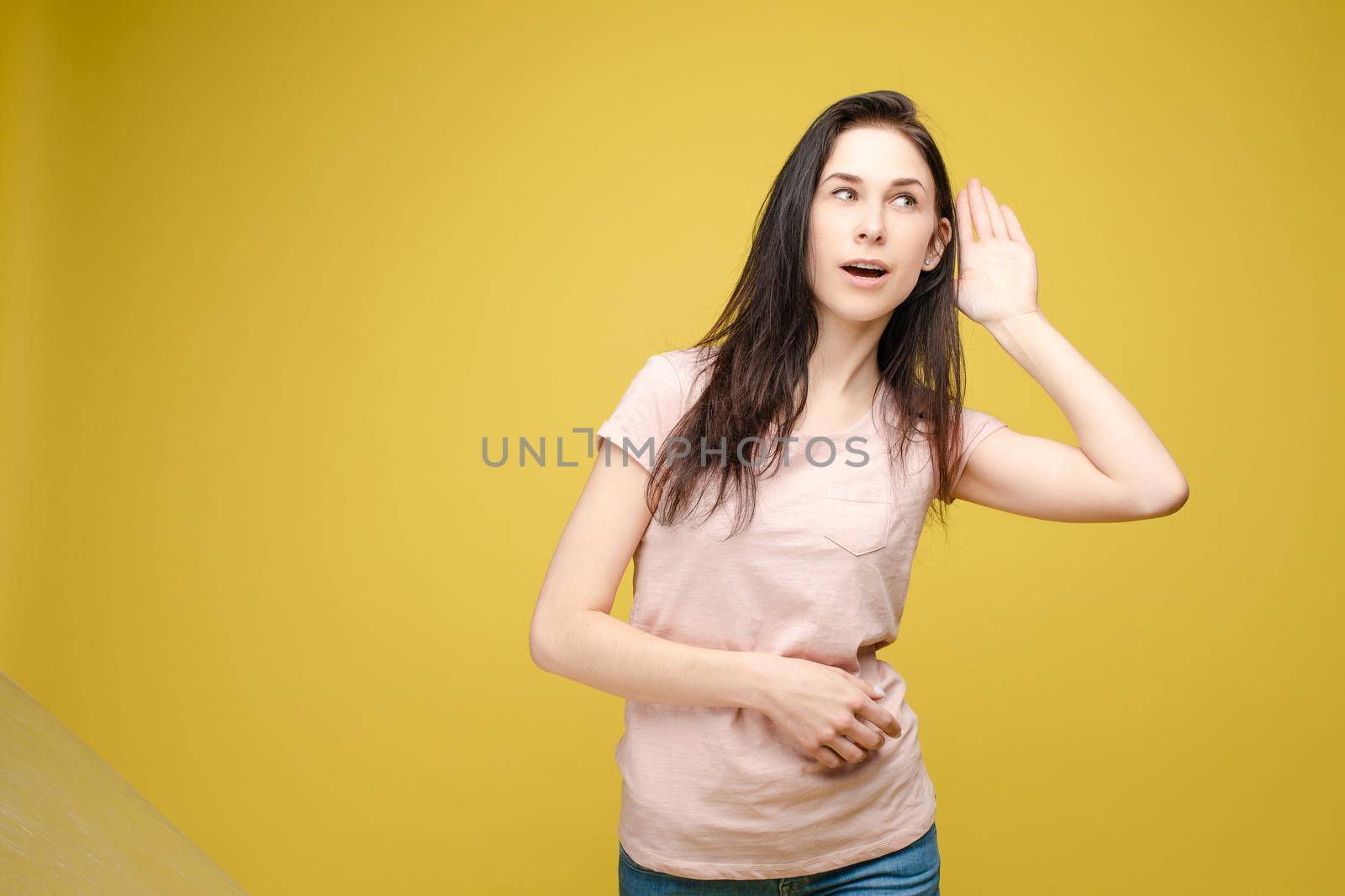 Studio portrait of curious brunette girl in multicolored top listening to the news or gossips with her ear.