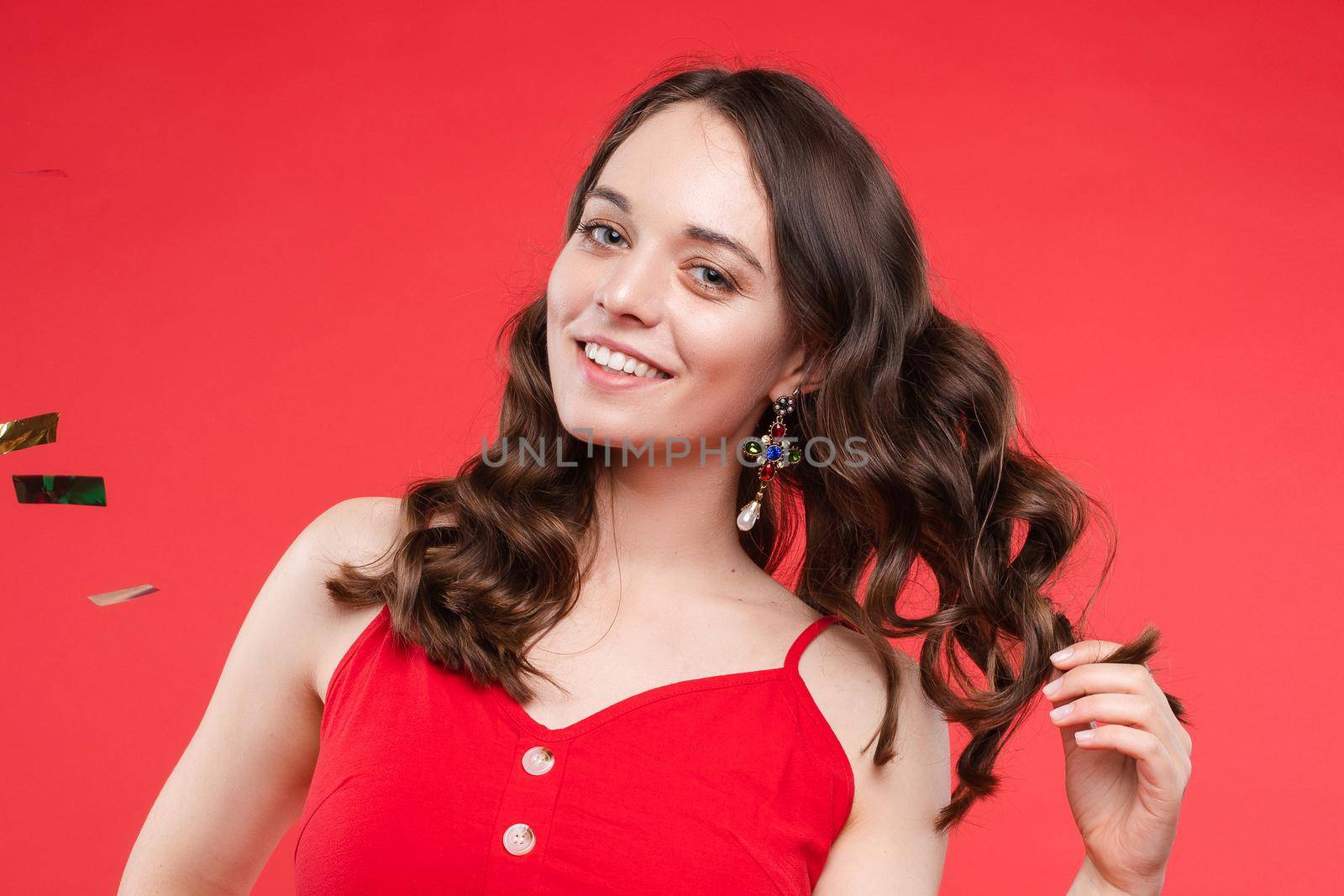 Front view of happy young girl wearing red dress looking at camera and twirling hair with hand in studio. Curly model posing and smiling on isolated background. Concept of emotions and celebration.