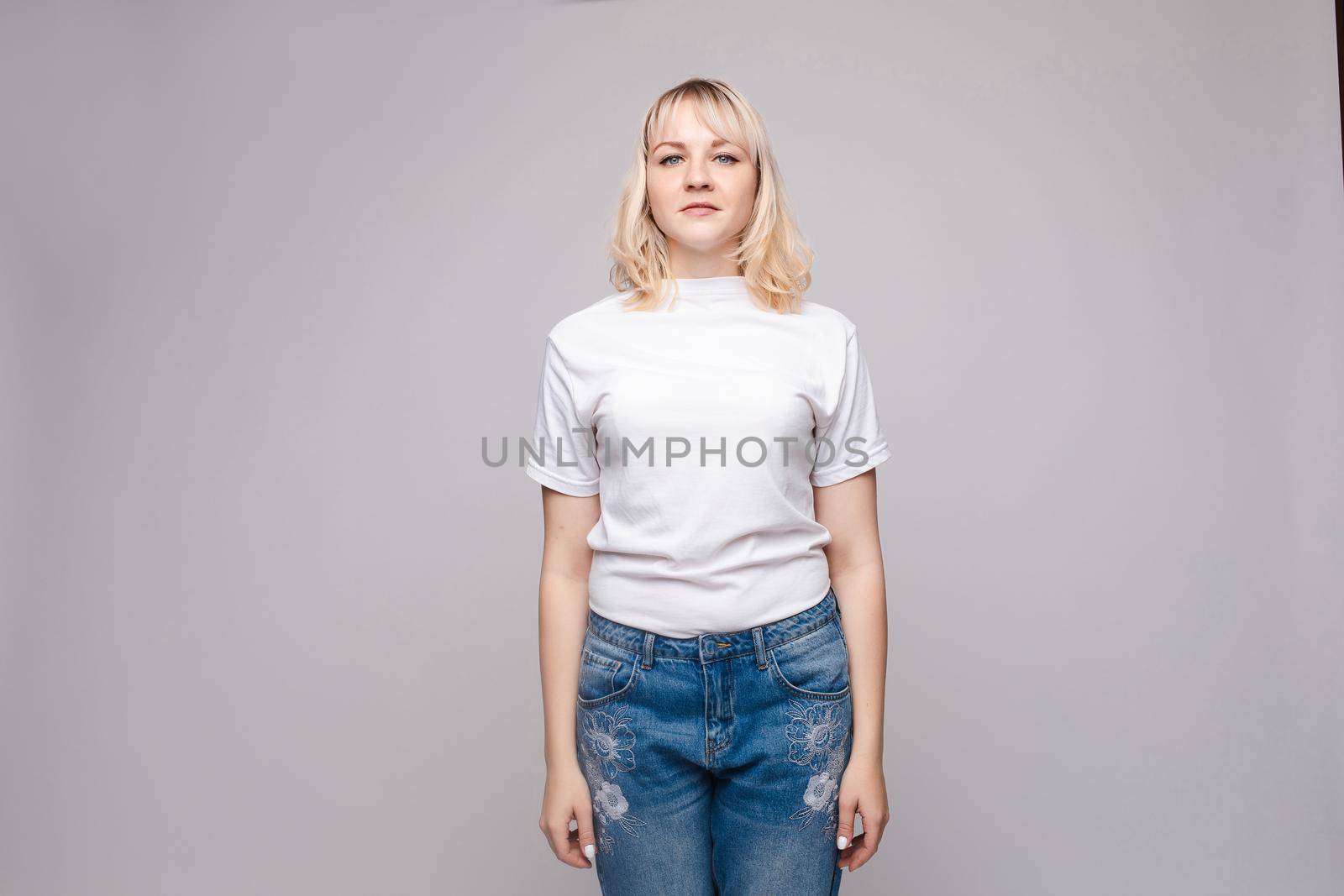 View from front of beautiful slim woman wearing white shirt and jeans standing steady on frey isolated background. Young blonde looking at camera, smiling and posing in studio.