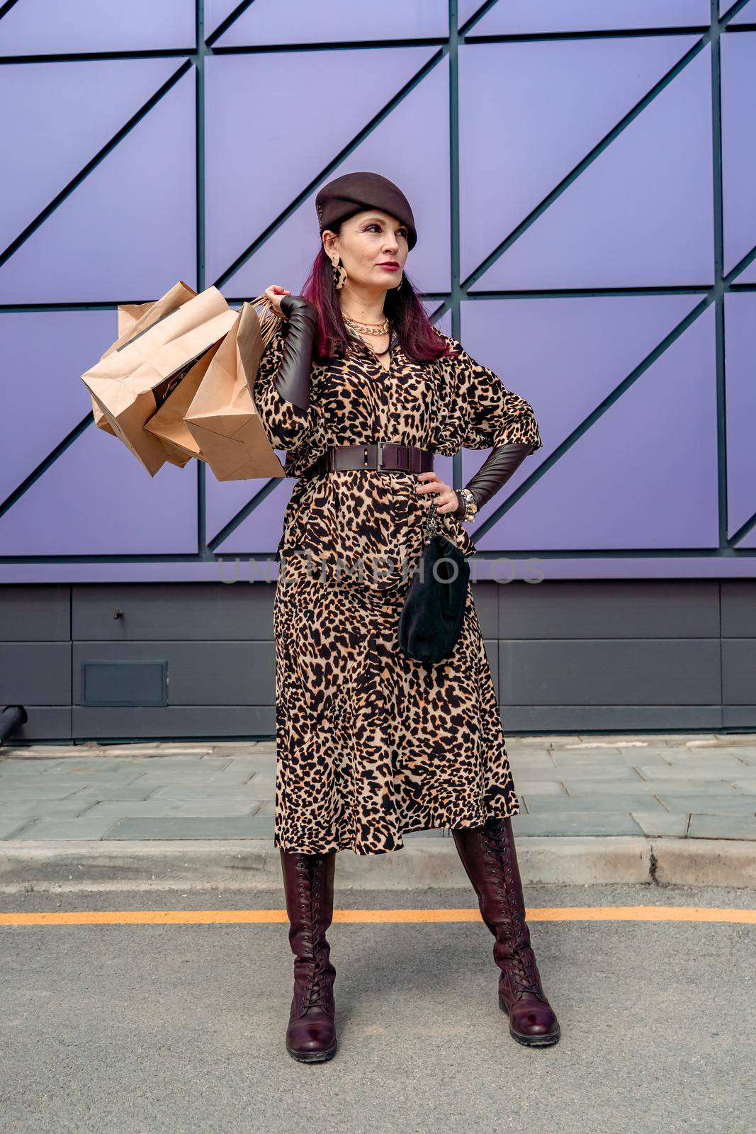 A happy shopaholic girl keeps her bags near the shopping center. A woman near the store is happy with her purchases, holding bags. Dressed in a leopard print dress. Consumer concept. by Matiunina