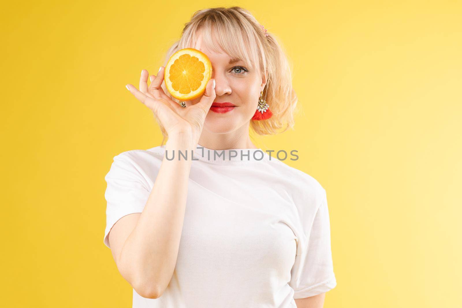 A woman with oranges in her eyes. Red lipstick lips. Merry, cheerful girl exudes positive, spoiled oranges. Isolated on yellow background by StudioLucky