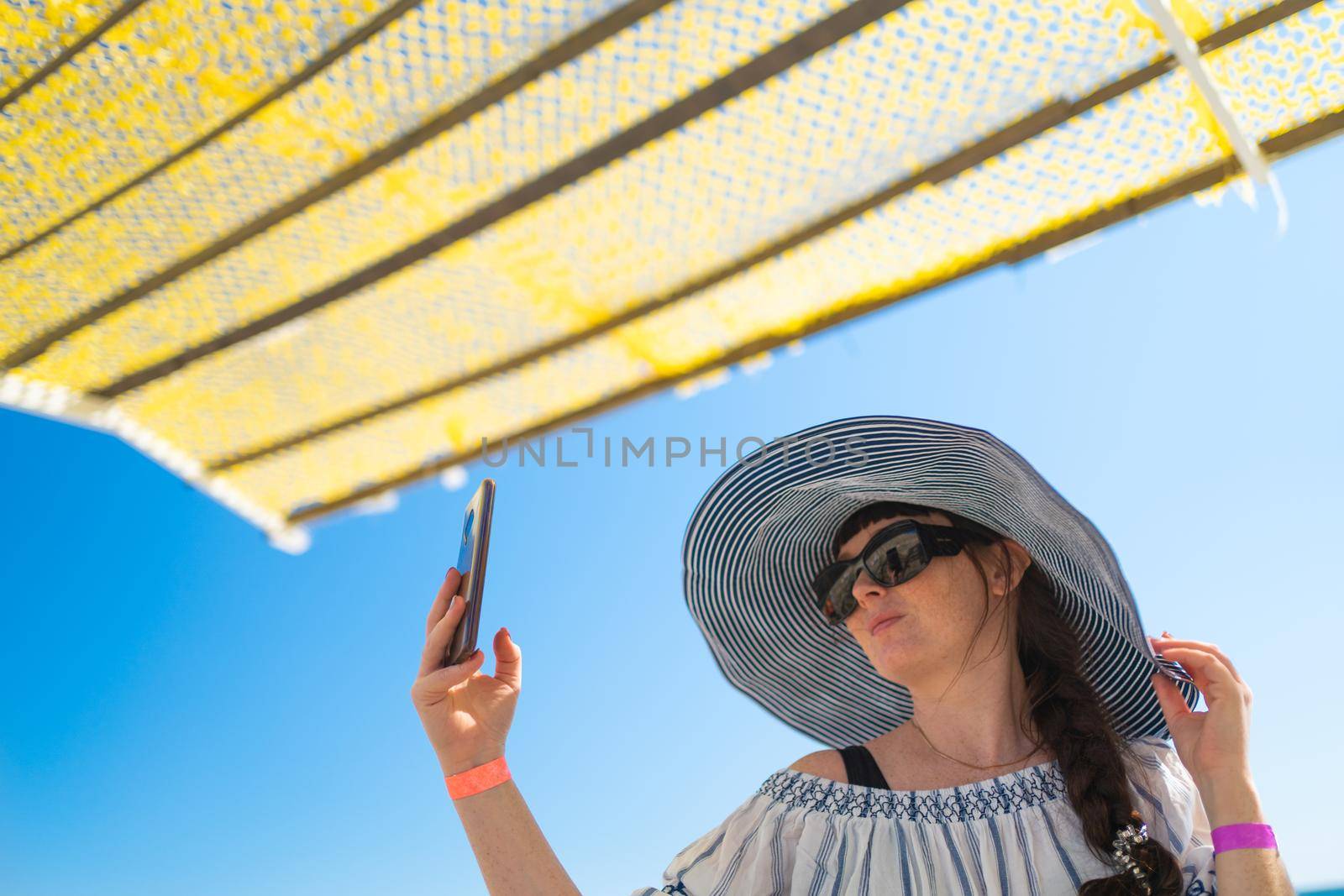 Summer beach holidays. on the beach, a girl in a striped hat with a large brim and a light cape under a yellow canopy protecting her from the scorching sun takes a selfie. The girl smiles and enjoys a sunny day. The concept of a happy lifestyle.