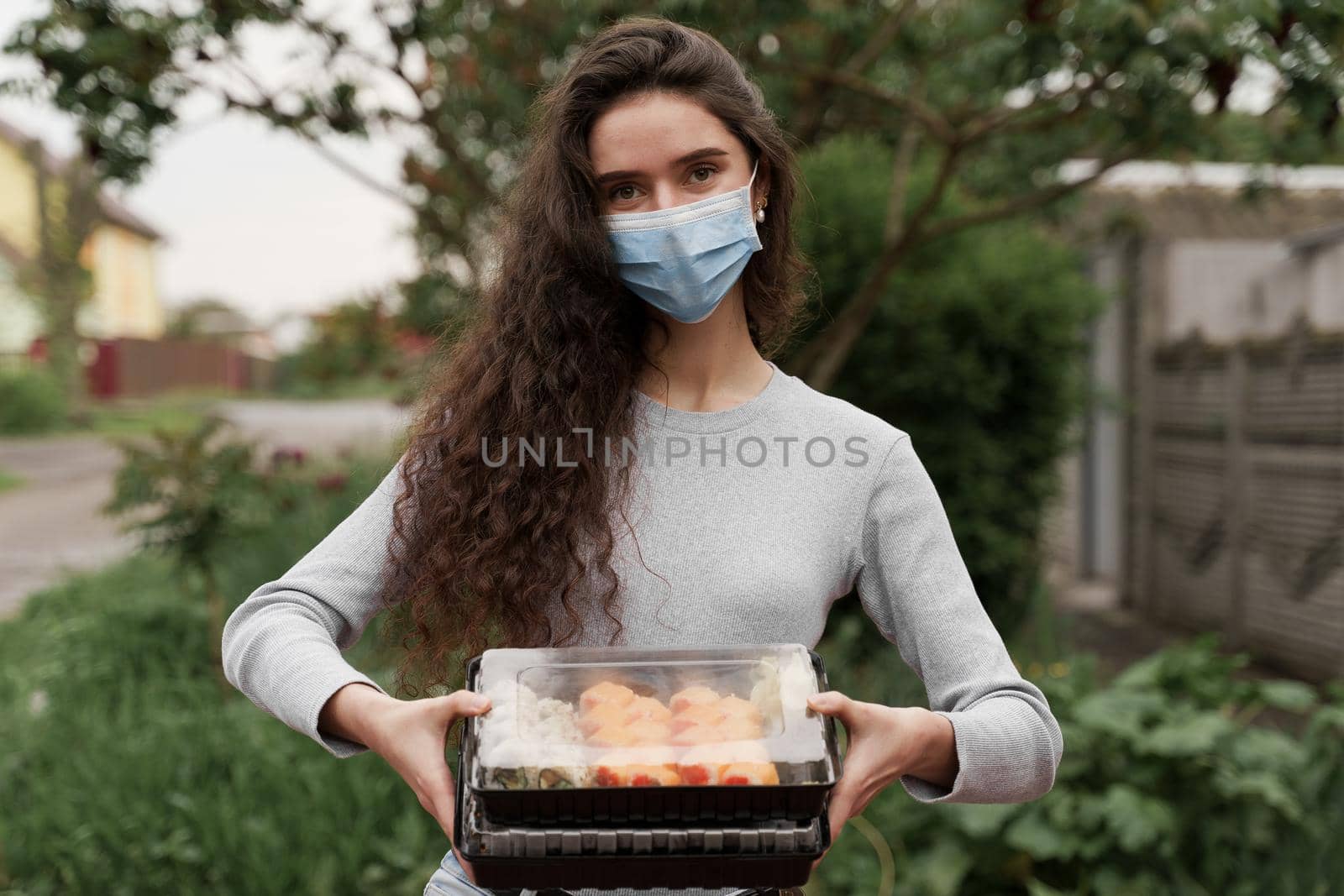 Sushi set in box healthy food delivery service by car. Girl courier in medical mask with 2 sushi boxes stands in front of car. by Rabizo