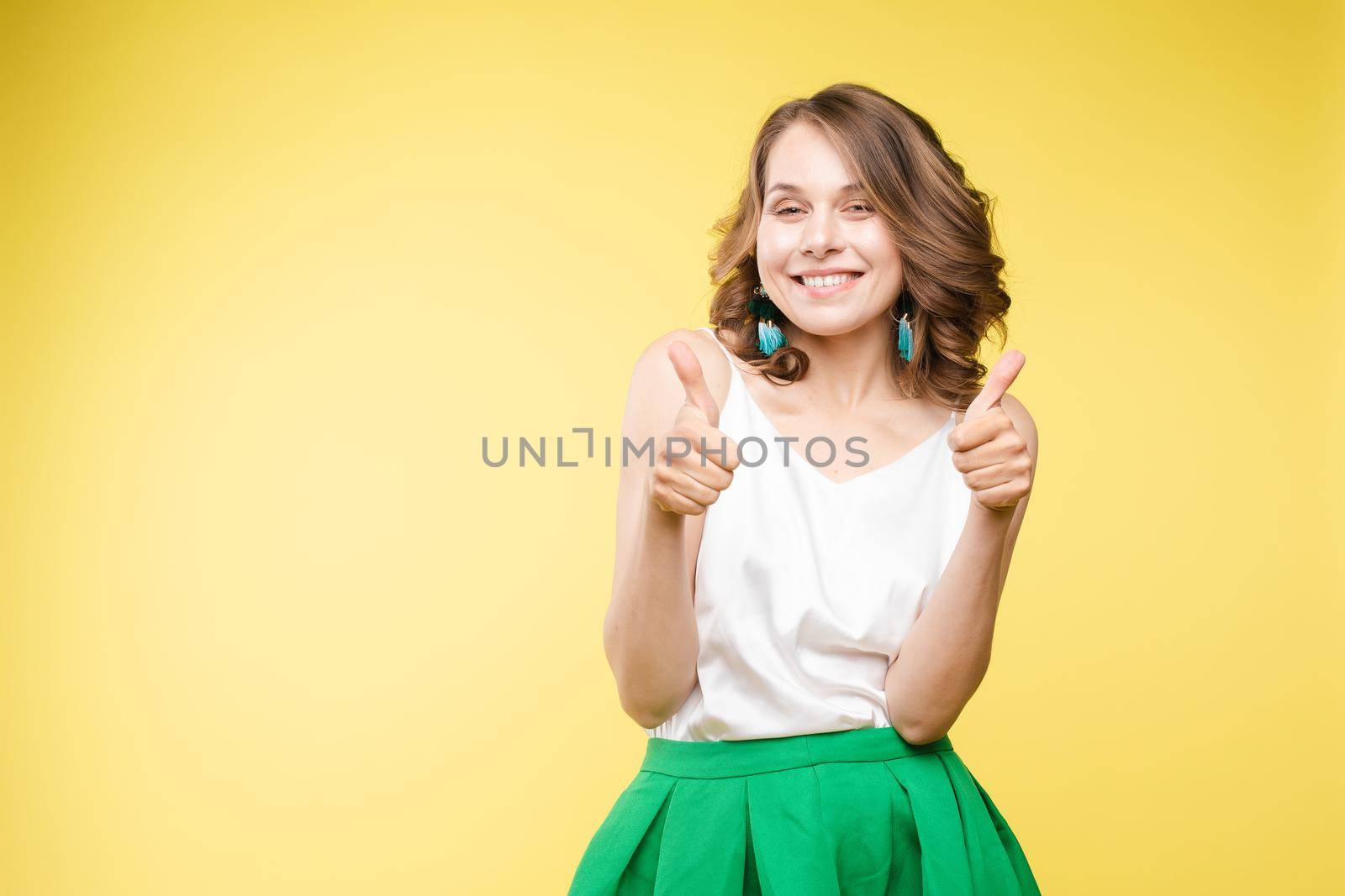 Front view of positive blonde wearing white shirt and jeans looking at camera and showing thumbs up. Young woman posing and laughing on grey isolated background. Concept of happiness.