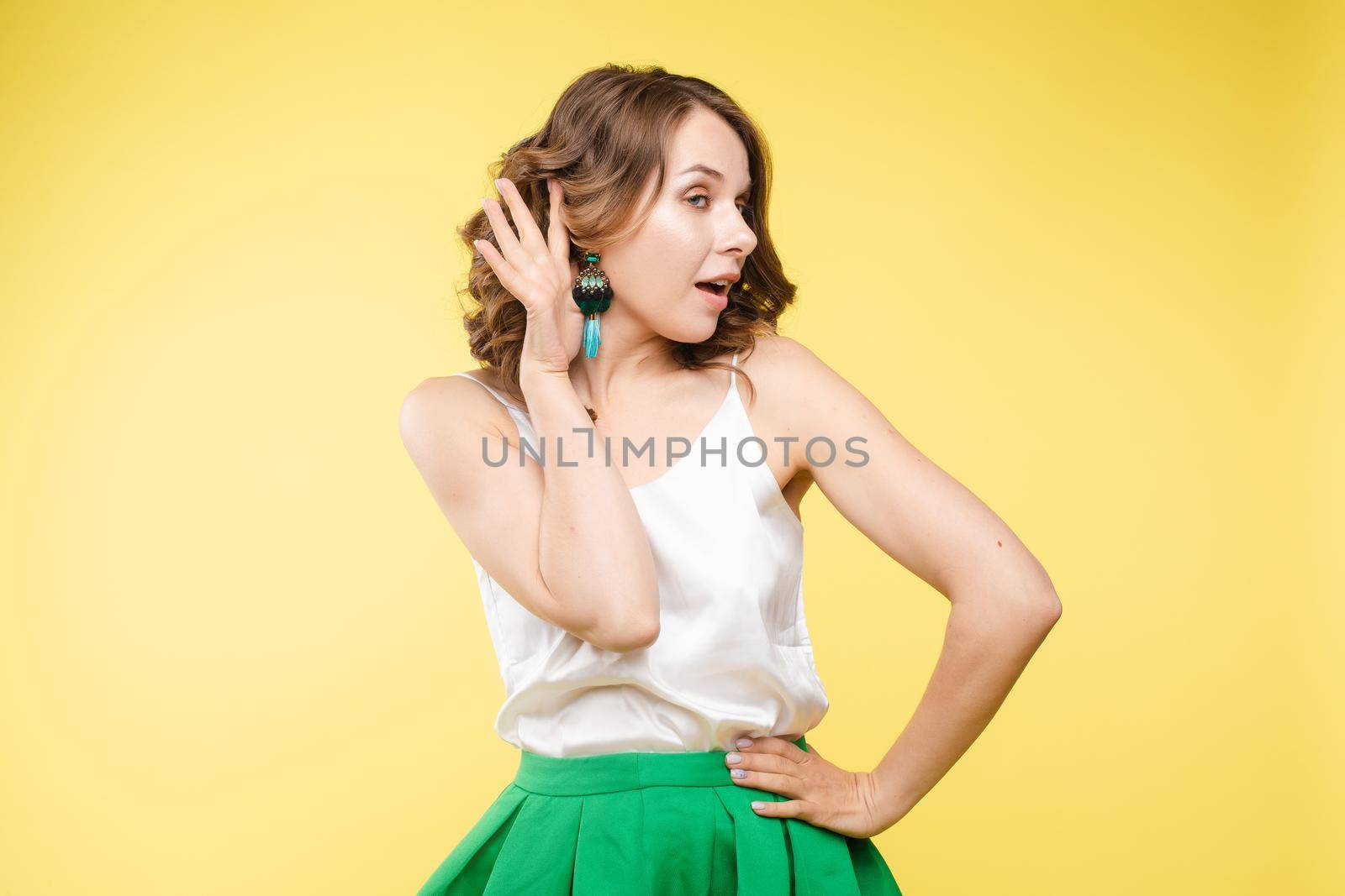 Studio portrait of curious brunette girl in multicolored top listening to the news or gossips with her ear.