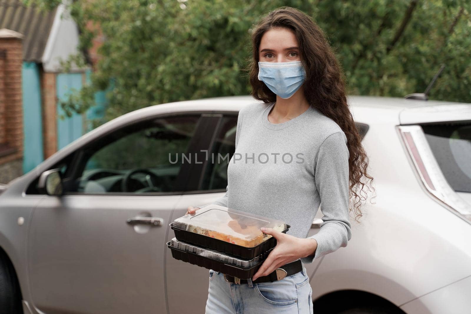 Sushi set in box healthy food delivery service by car. Girl courier in medical mask with 2 sushi boxes stands in front of car. by Rabizo