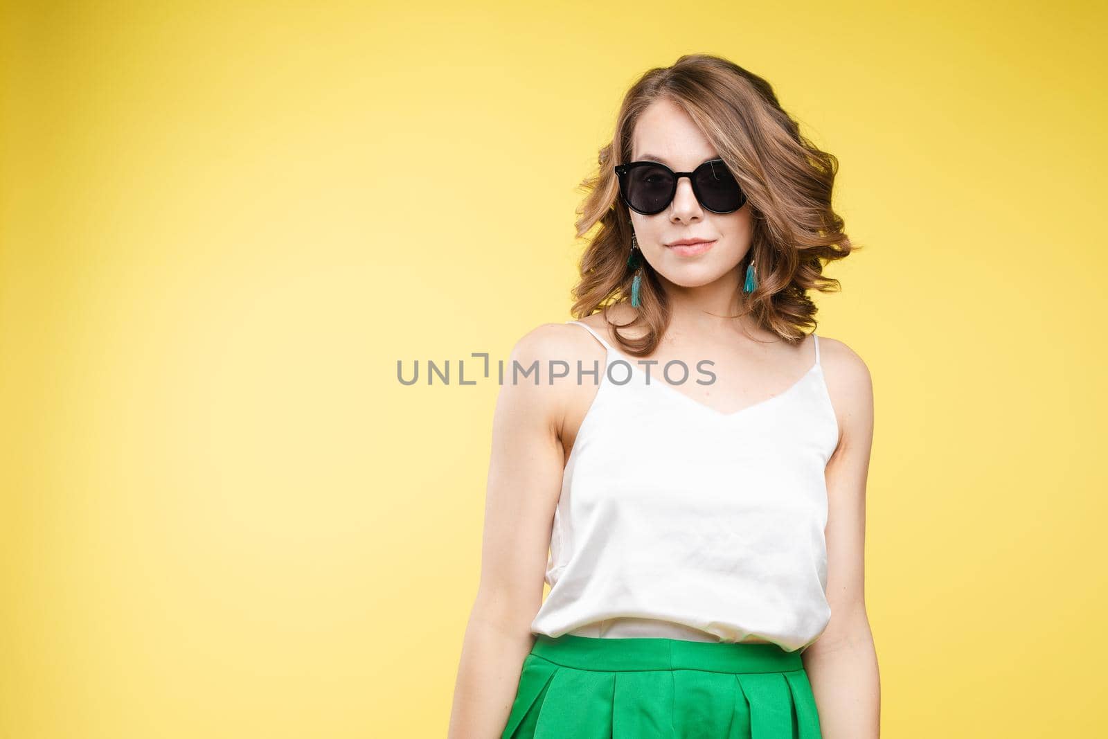 Summer outfit on orange background. Stylish and trendy beautiful young girl with wavy middle-length hair in white top and green bottom wearing trendy earrings and black sunglasses. She is smiling over orange background. Copyspace. Isolate.