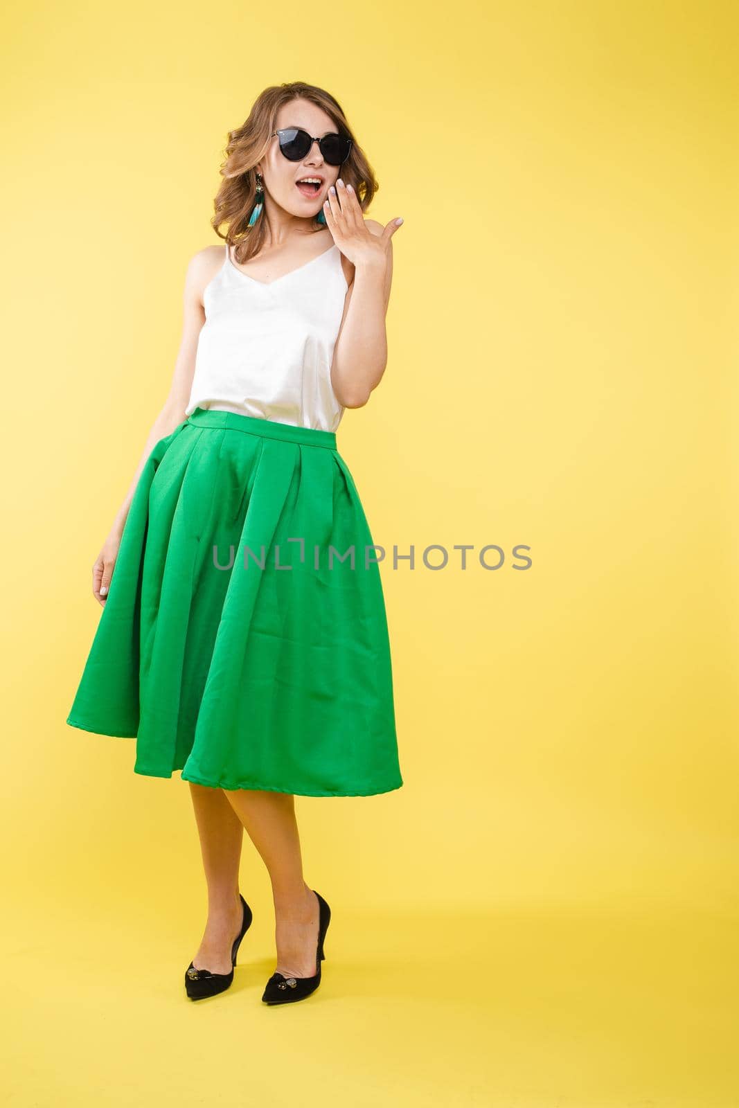 Summer outfit on orange background. Stylish and trendy beautiful young girl with wavy middle-length hair in white top and green bottom wearing trendy earrings and black sunglasses. She is smiling over orange background. Copyspace. Isolate.