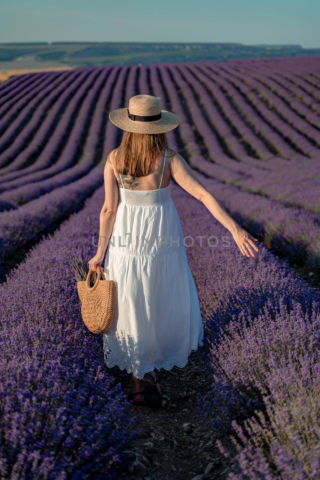 A middle-aged woman, blonde in a white dress and hat, walks through a lavender field with a basket.