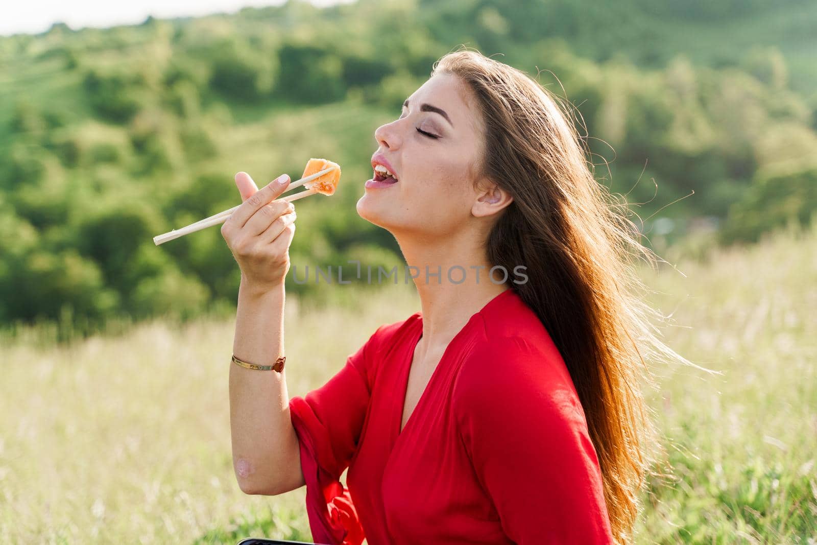 Sushi set and girl who reach out to bite sushi on green hills background. Food delivery service from japanese restaurant. Woman with blue eyes in red dress seats and eats sushi delivered by courier