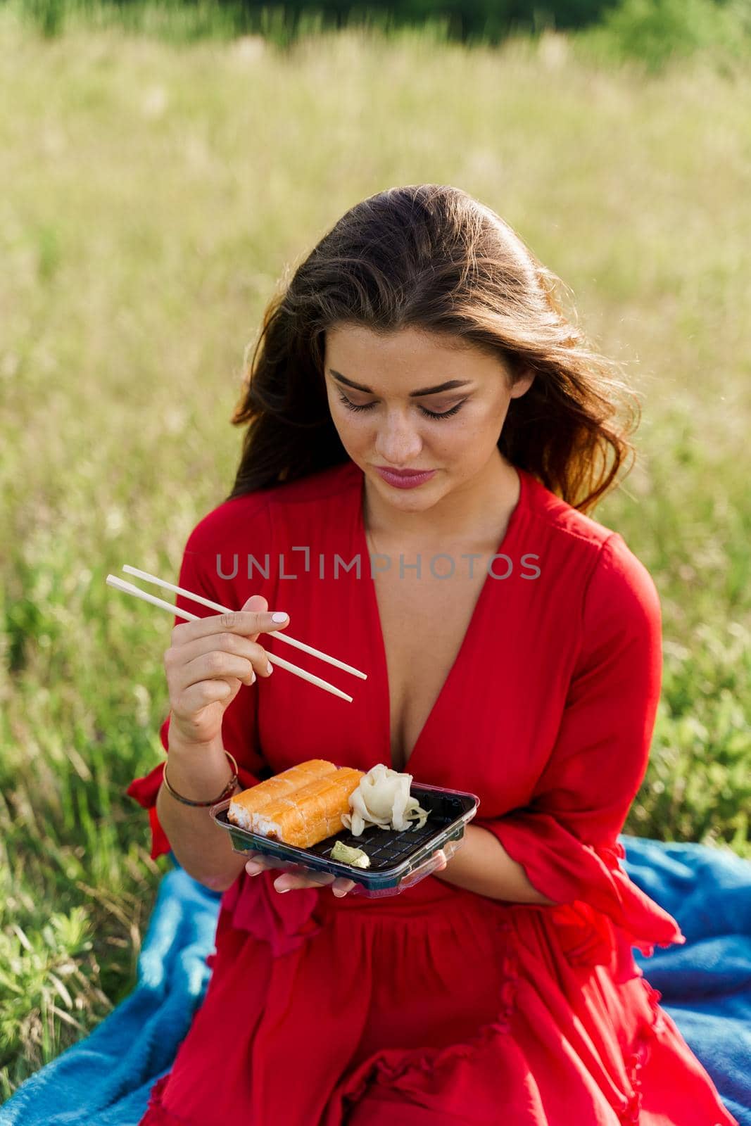 Delivery sushi set in box for girl. Attractive girls in red dress seats in green park and eat sushi. Vertical photo for social networks advert for restaurant. by Rabizo