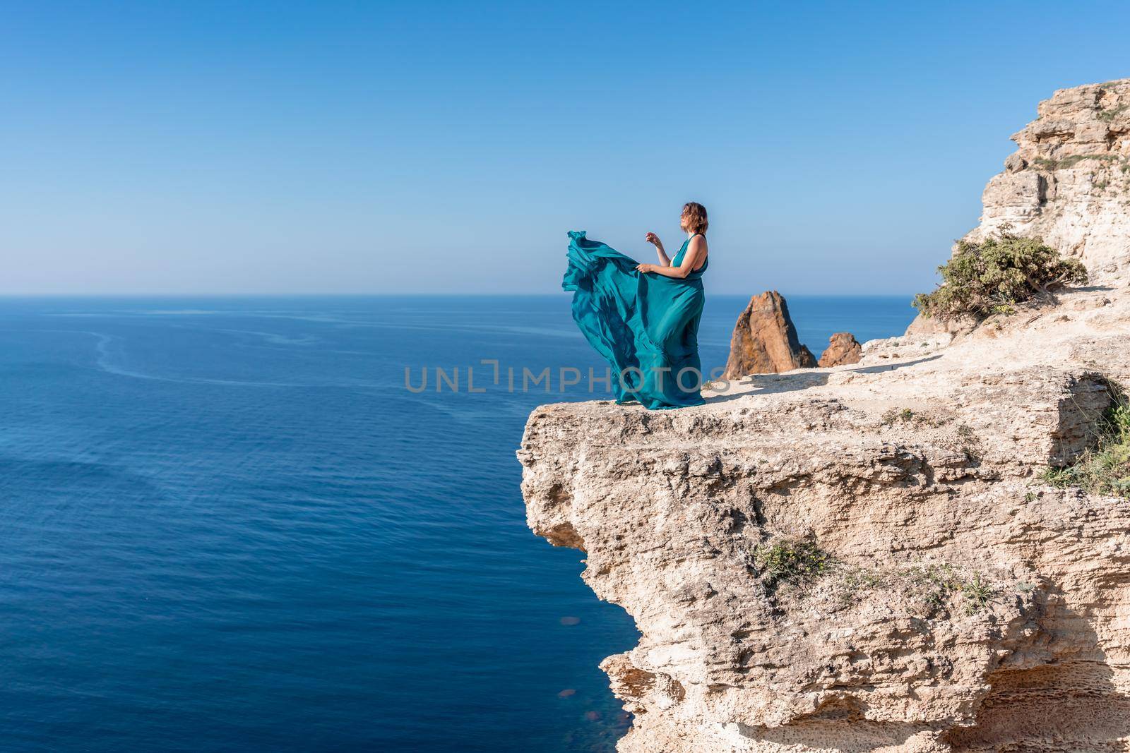 A girl with loose hair in a long mint dress descends the stairs between the yellow rocks overlooking the sea. A rock can be seen in the sea. Sunny path on the sea from the rising sun.