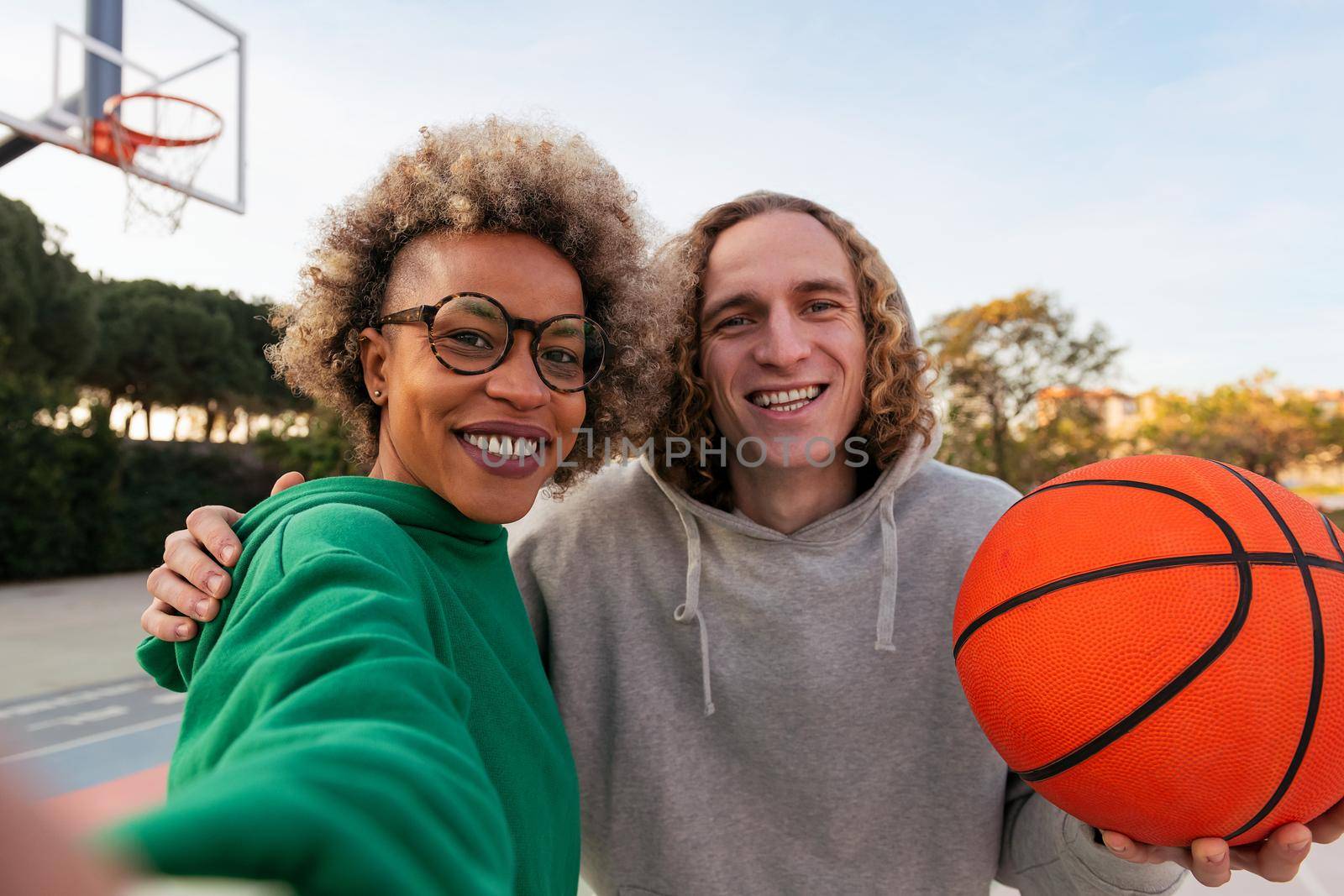 selfie of a latin woman and caucasian man smiling happy after playing in a city park basketball court, concept of friendship and urban sport in the street