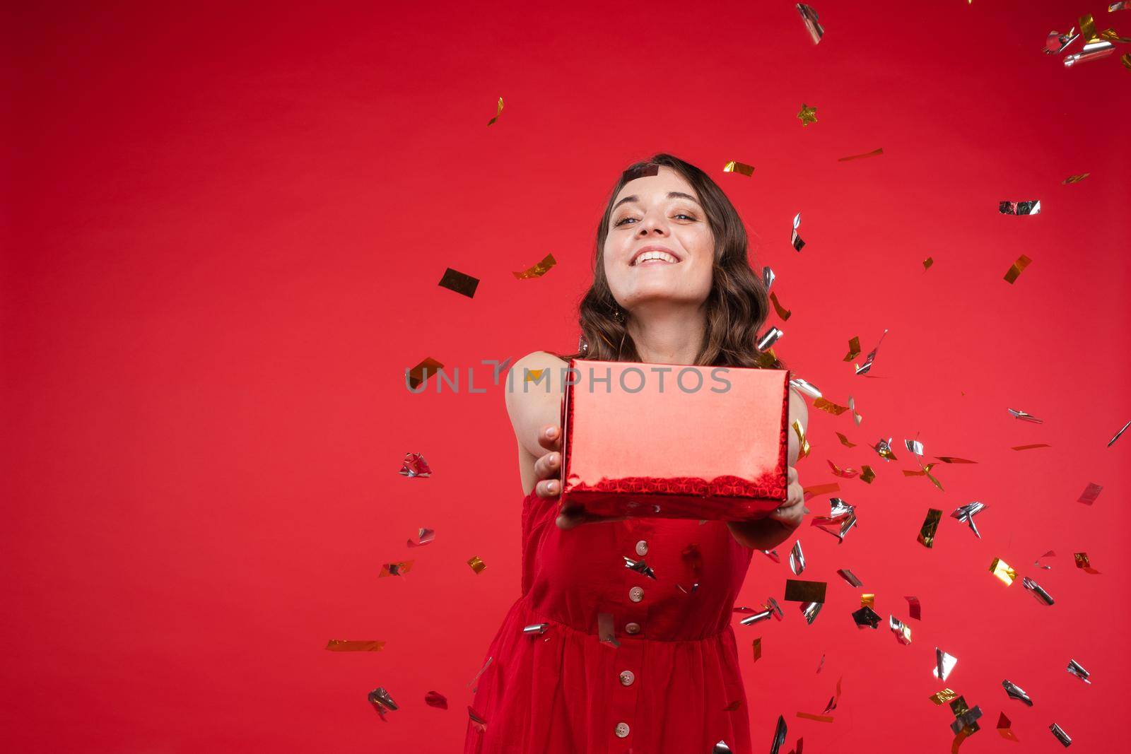 Studio portrait of beautiful young brunette lady with wavy hair in summer red dress with buttons smiling at camera. She is sprinkled or showered with sparkling confetti on red background. Isolate.