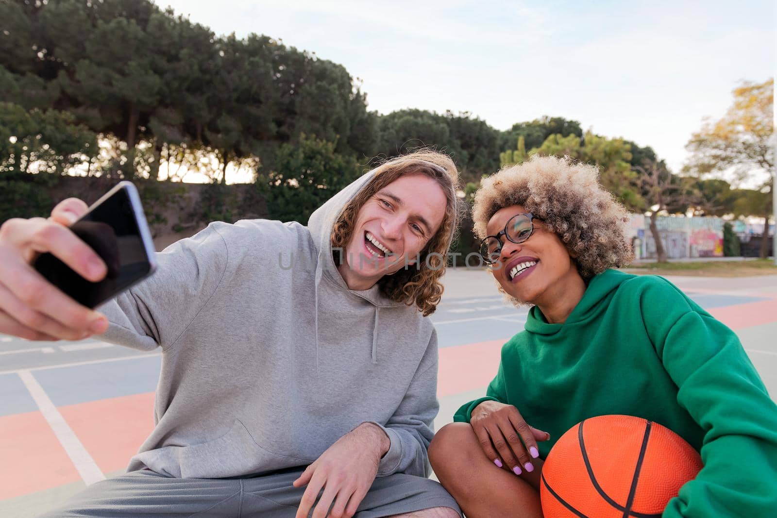 man and woman sharing a good time taking a selfie with the phone after playing basketball in a city park, concept of friendship and urban sport in the street, copy space for text