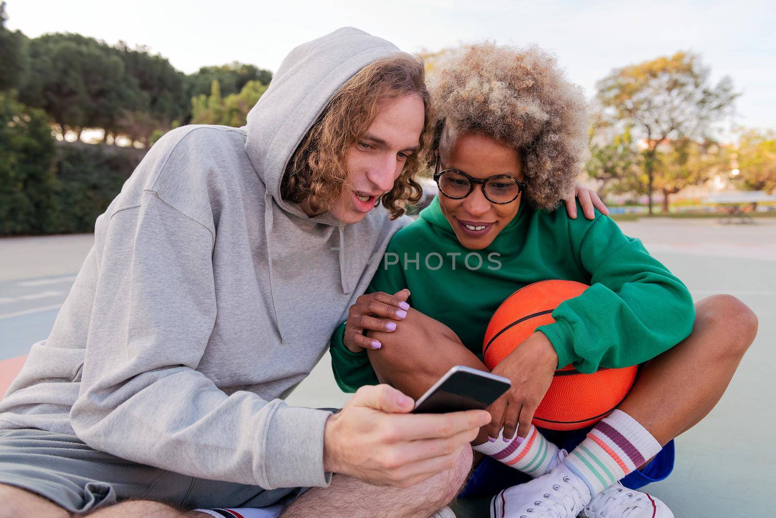 man and woman have a good time looking at things on the cell phone while sitting after basketball practice in a city park, concept of friendship and urban sport in the street