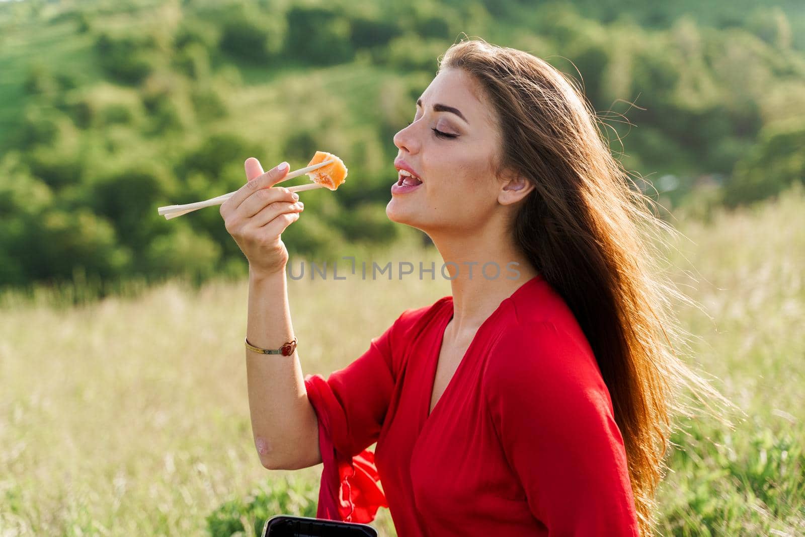 Sushi set and girl who reach out to bite sushi on green hills background. Food delivery service from japanese restaurant. Woman with blue eyes in red dress seats and eats sushi delivered by courier
