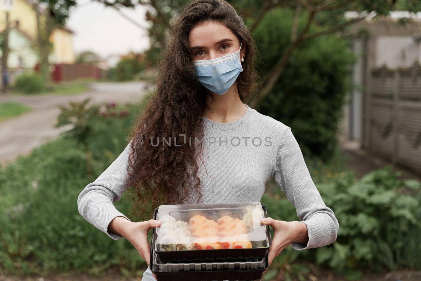 Girl courier in medical mask with 2 sushi boxes stands in front of car. Sushi set in box healthy food delivery service by car