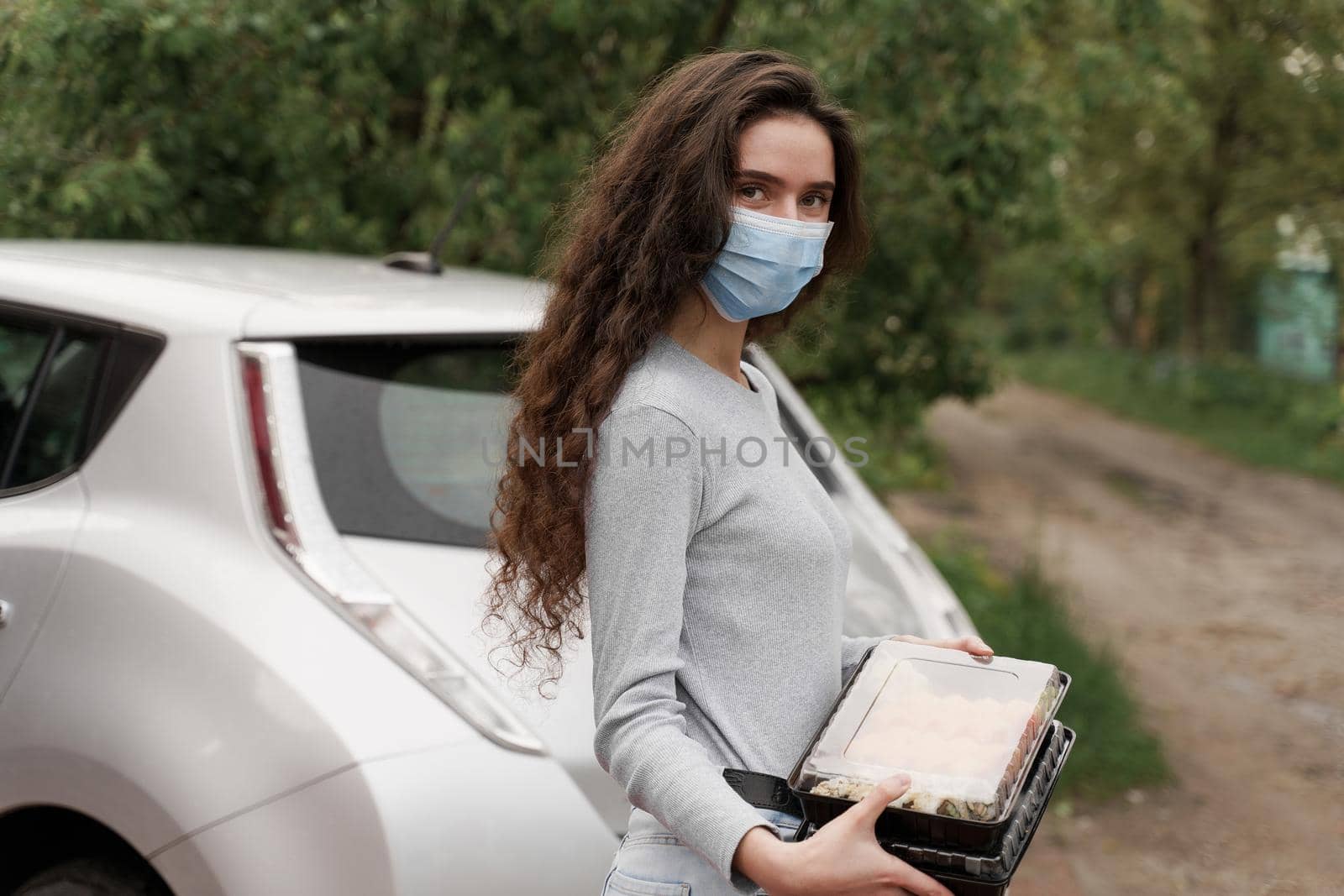 Sushi set in box healthy food delivery service by car. Girl courier in medical mask with 2 sushi boxes stands in front of car