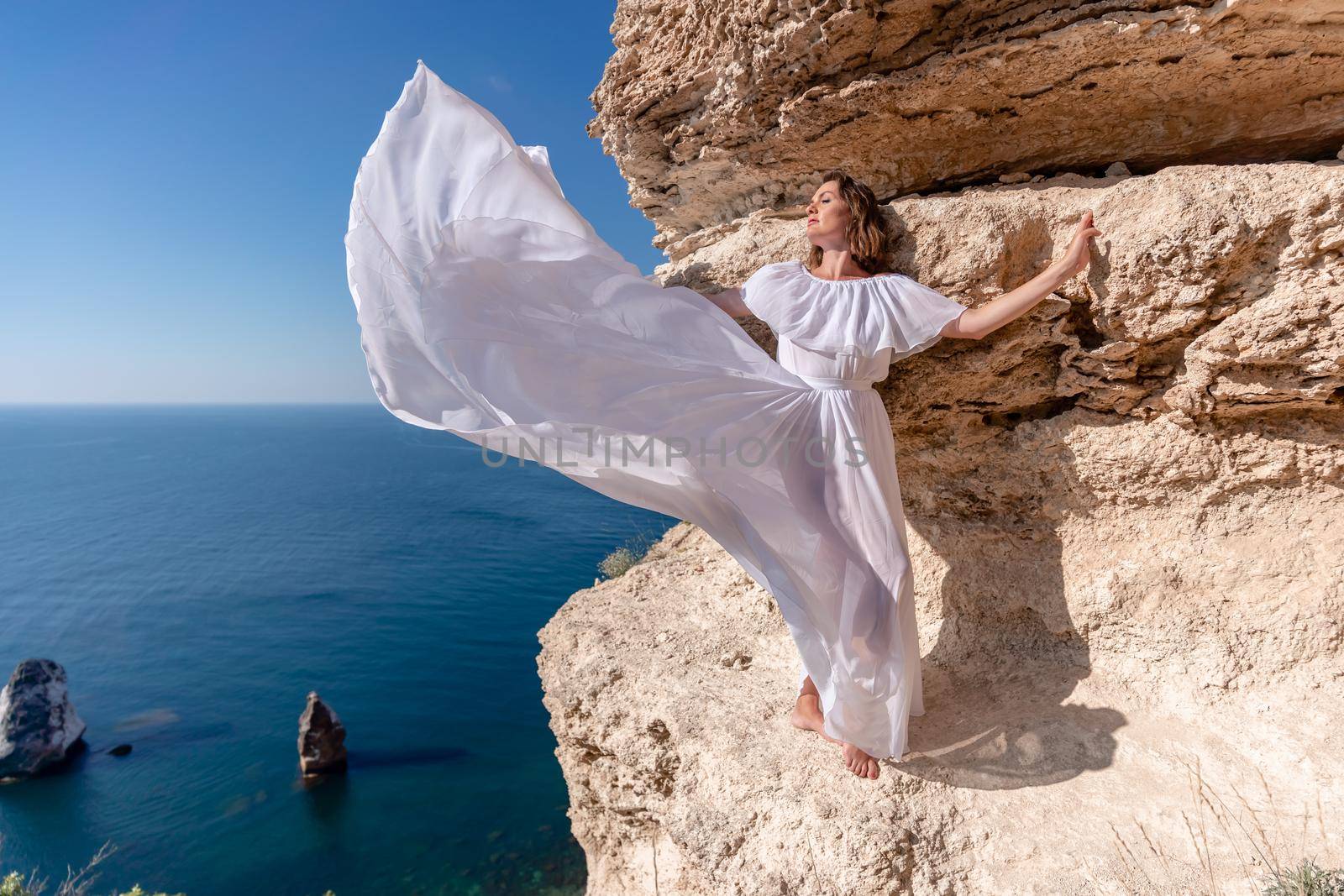 A beautiful young woman in a white light dress with long legs stands on the edge of a cliff above the sea waving a white long dress, against the background of the blue sky and the sea