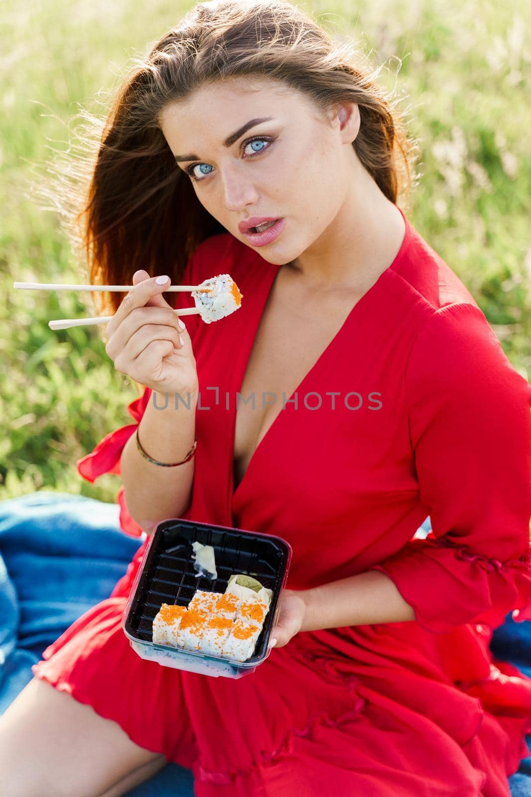 Delivery sushi set in box for girl. Attractive girls in red dress seats in green park and eat sushi. Vertical photo for social networks advert for restaurant