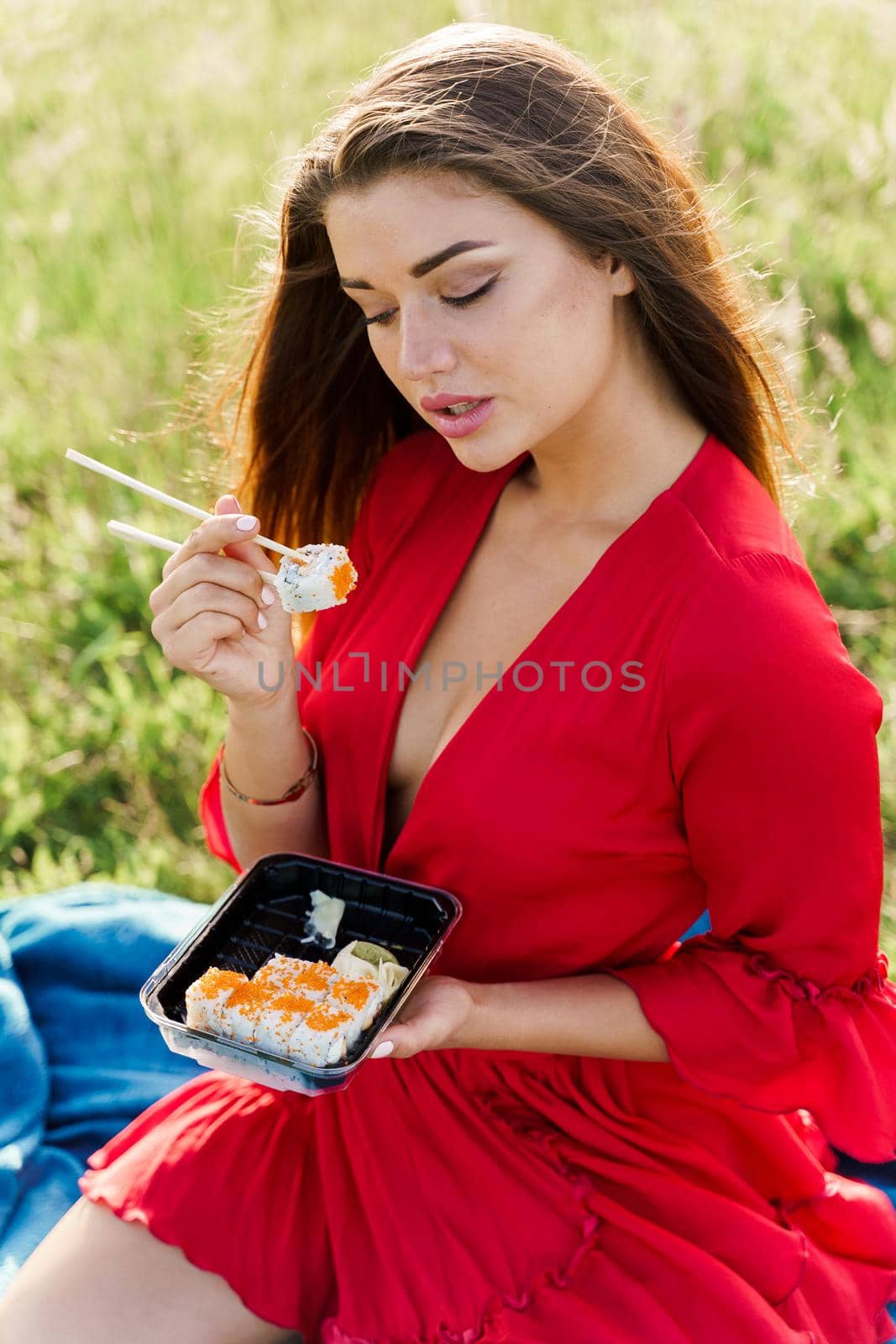 Delivery sushi set in box for girl. Attractive girls in red dress seats in green park and eat sushi. Vertical photo for social networks advert for restaurant