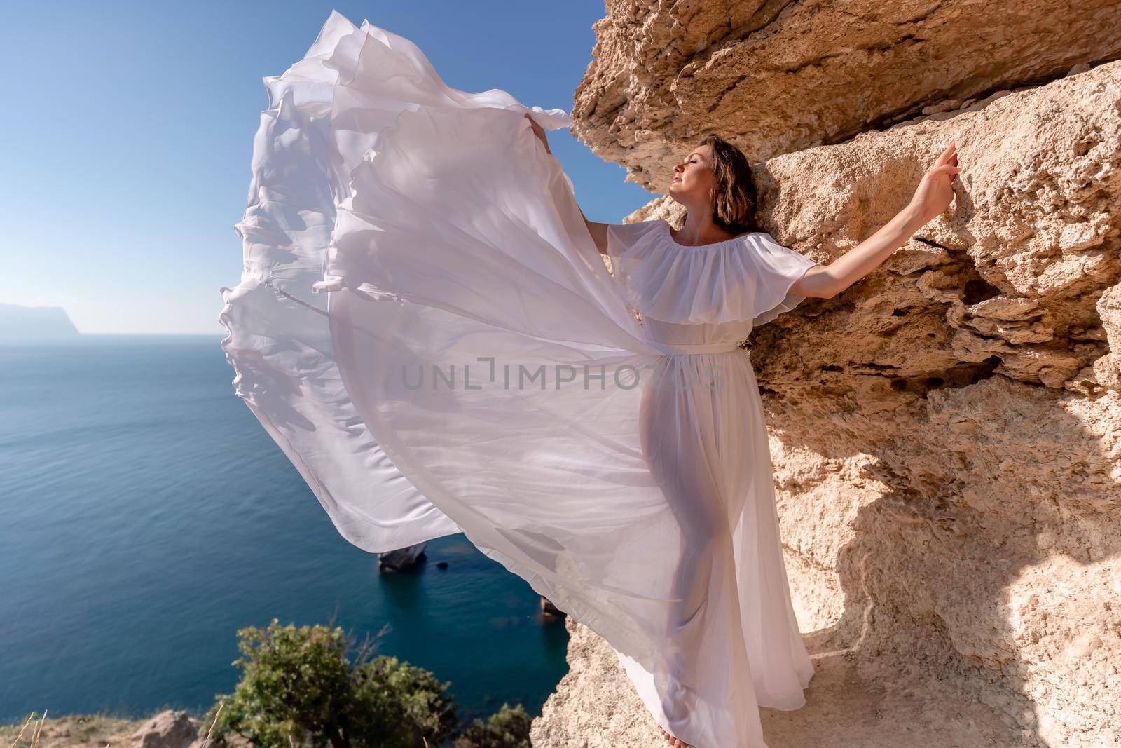 A beautiful young woman in a white light dress with long legs stands on the edge of a cliff above the sea waving a white long dress, against the background of the blue sky and the sea