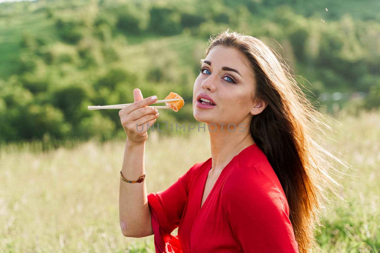 Sushi set and girl who reach out to bite sushi on green hills background. Food delivery service from japanese restaurant. Woman with blue eyes in red dress seats and eats sushi delivered by courier
