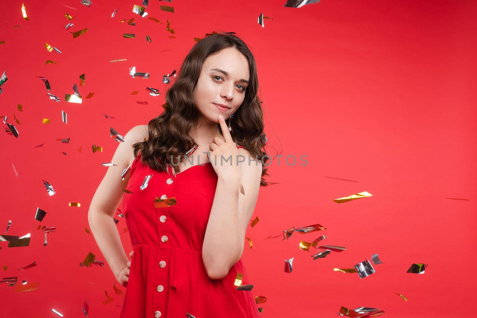Front view of happy young girl wearing red dress looking at camera and twirling hair with hand in studio. Curly model posing and smiling on isolated background. Concept of emotions and celebration.