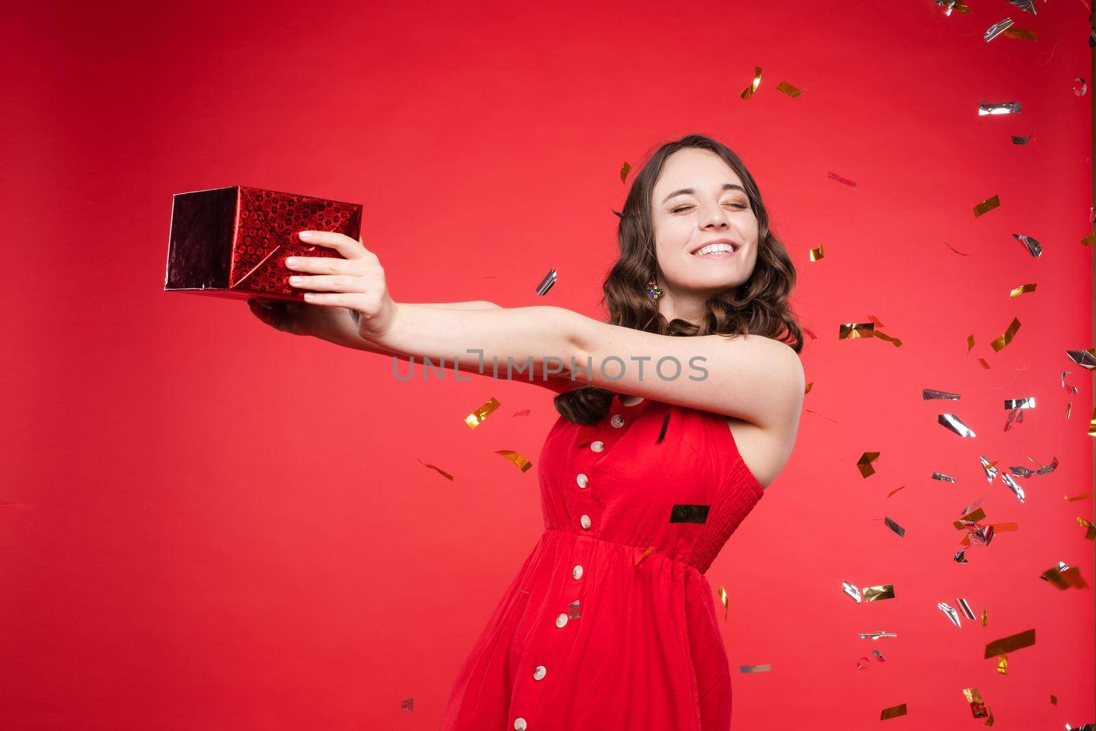 Studio portrait of beautiful young brunette lady with wavy hair in summer red dress with buttons smiling at camera. She is sprinkled or showered with sparkling confetti on red background. Isolate.