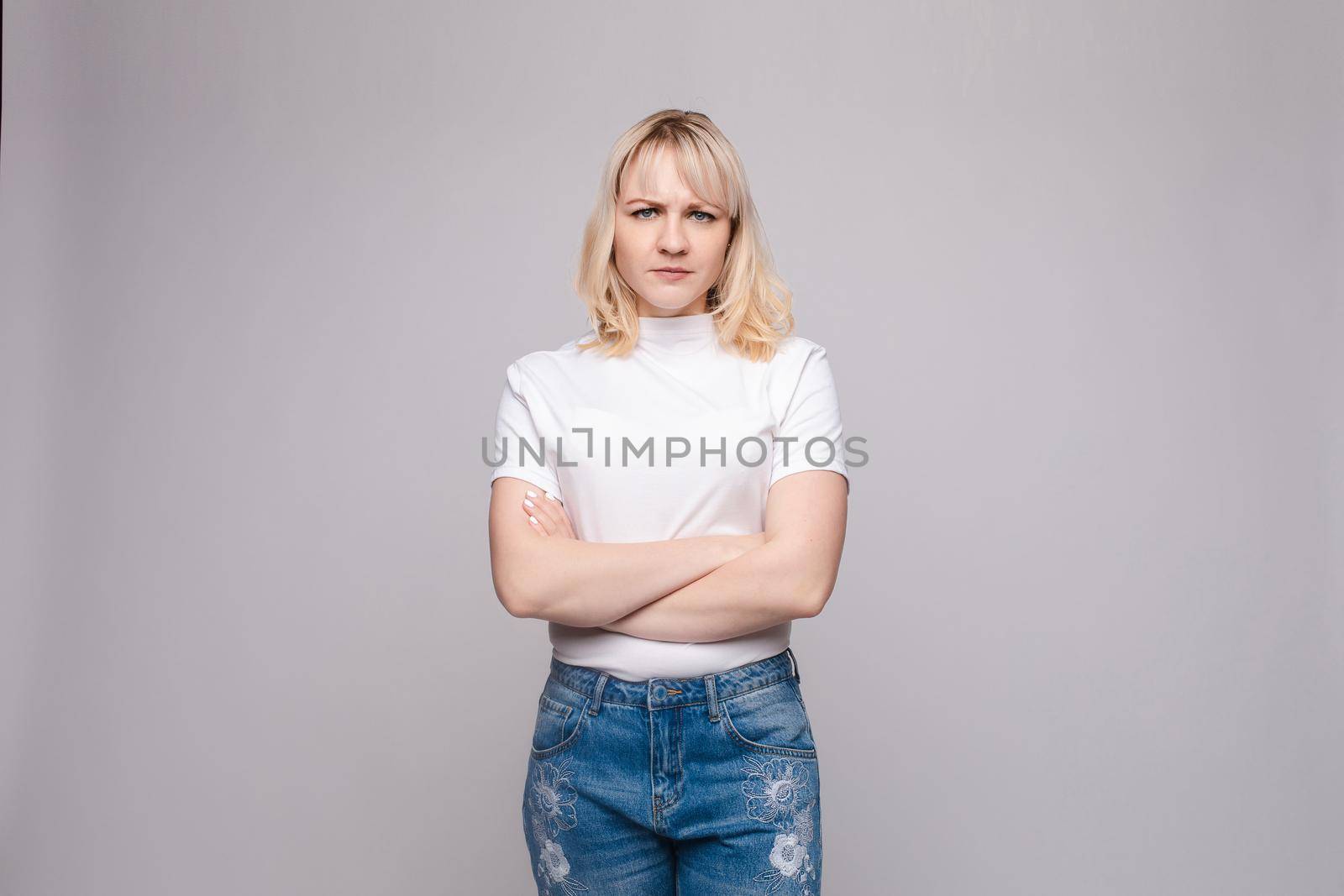 Studio portrait of unsatisfied young brunette caucasian woman with wavy hair in overall with colorful pattern holding arms folded and looking at camera with grief, dissatisfaction, anger and disbelief. Isolate.