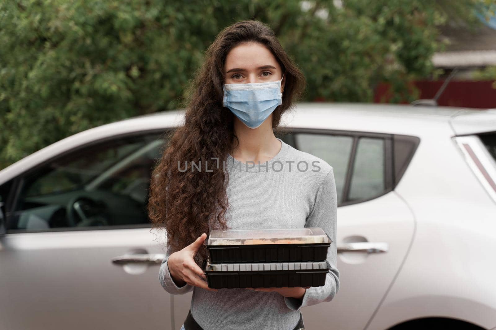 Sushi set in box healthy food delivery service by car. Attractive girl in medical mask with 2 sushi boxes stands in front of car. by Rabizo